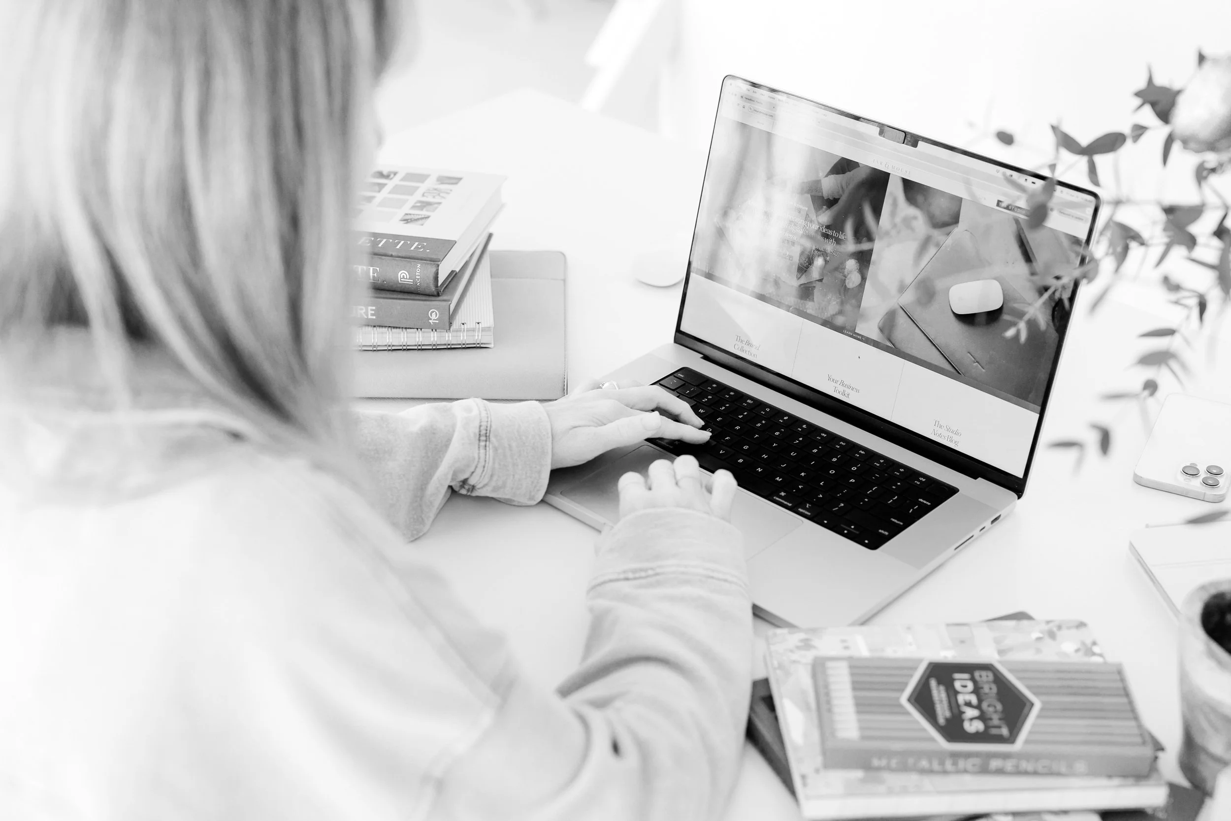 A person using a laptop on a desk with books, notebooks, a mouse, and a smartphone.