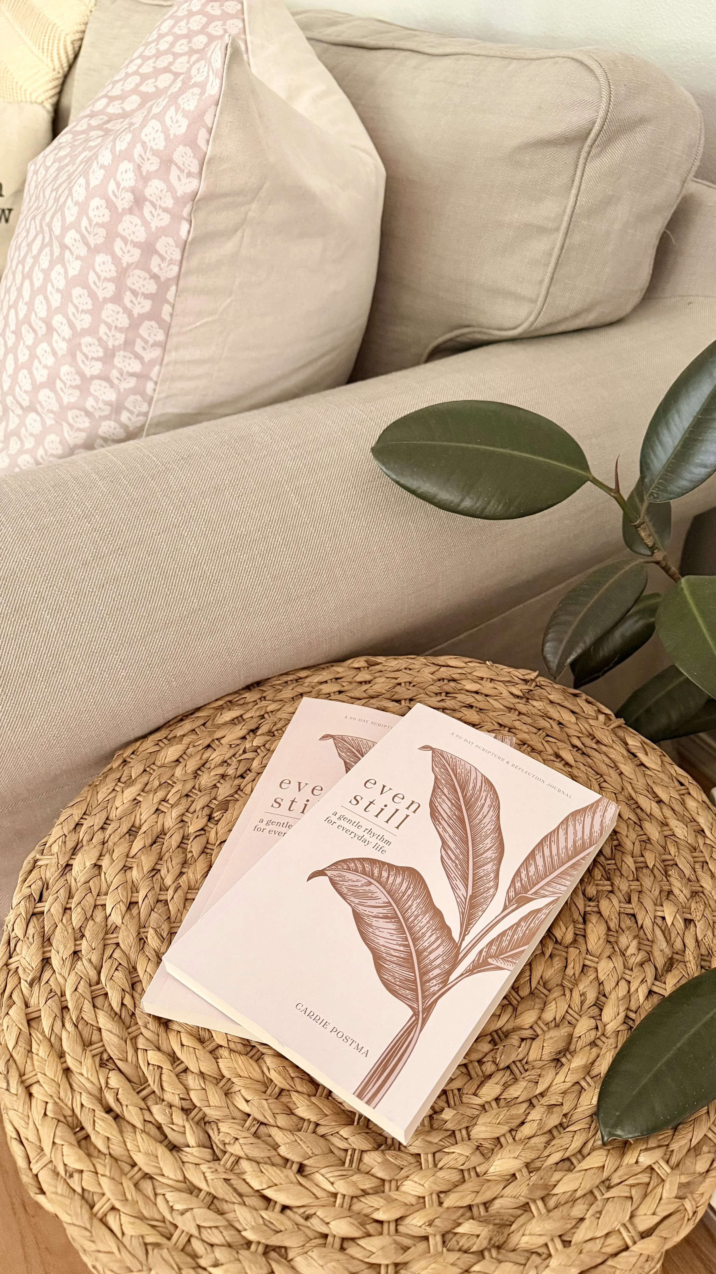 A woven rattan coffee table with three booklets titled 'Even Still' featuring a leaf illustration, next to a beige sofa with decorative pillows and a green leafy plant.
