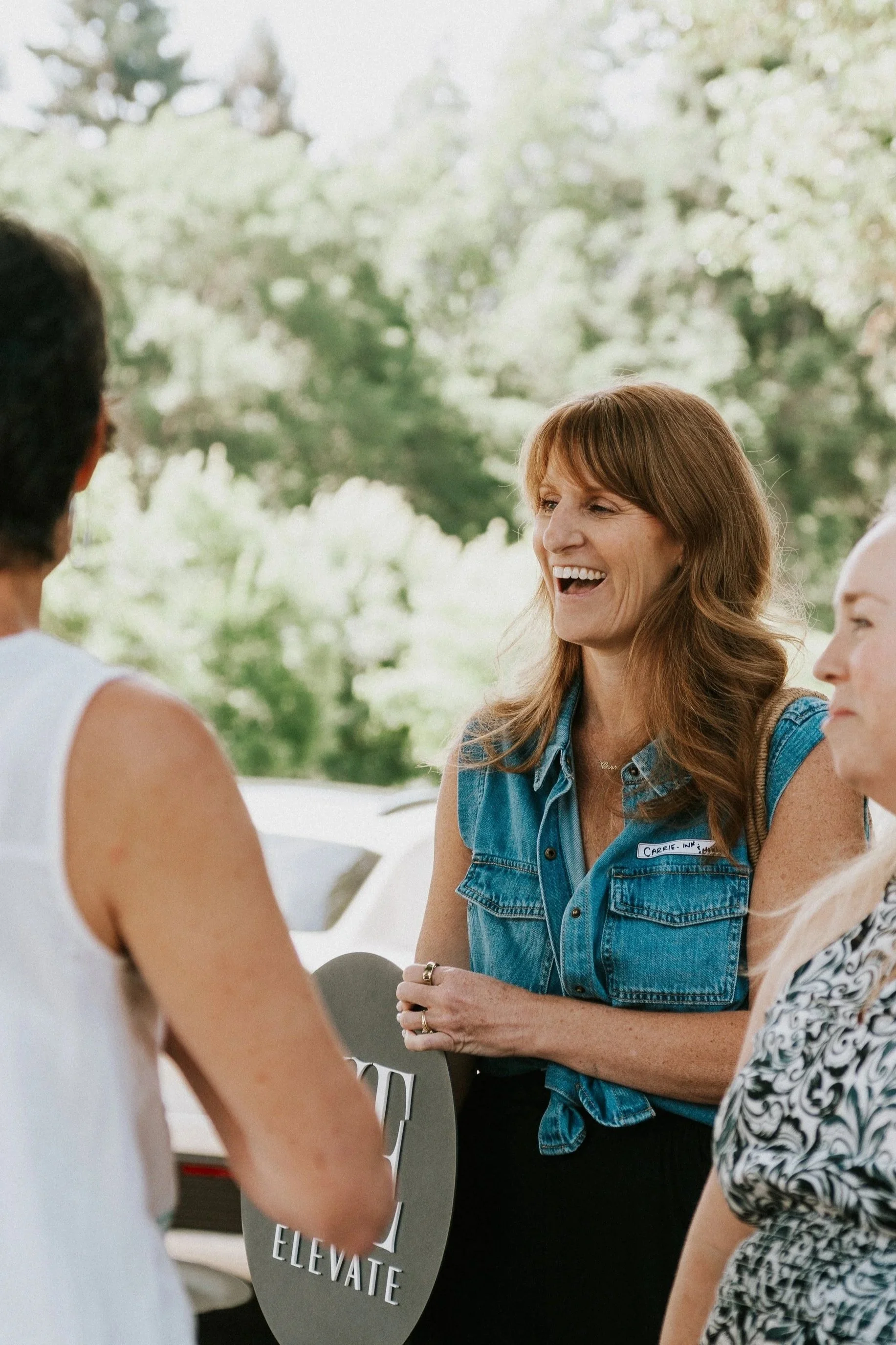 Three women talking outdoors, one holding a sign that says 'ELEVATE', with trees in the background.