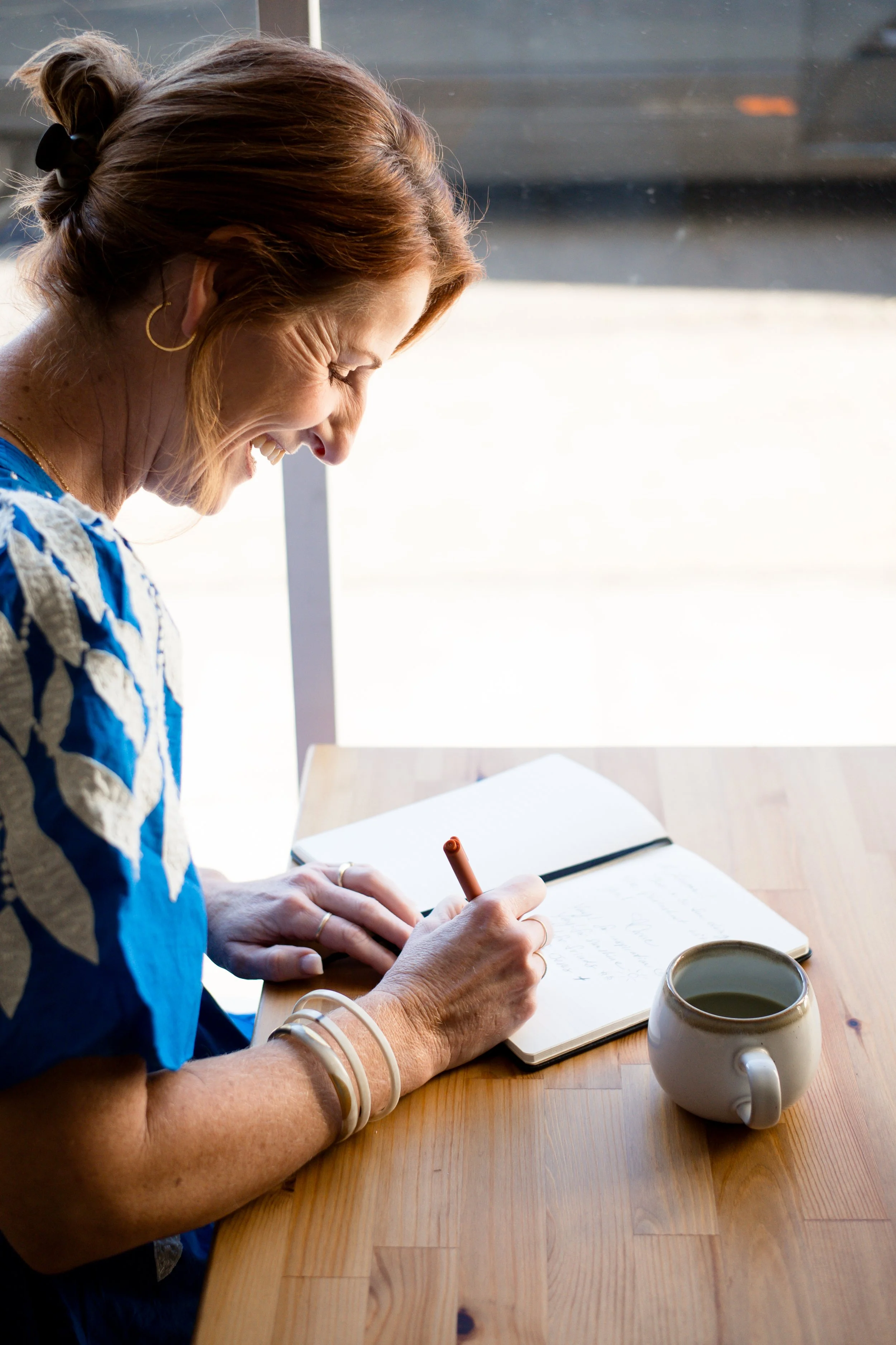 A woman with light skin and reddish hair tied back, wearing hoop earrings, a blue patterned top, and jewelry on her right arm, is smiling while writing in a notebook at a wooden table. There are two cups on the table, one of which is filled with a dark beverage. She is sitting near a window with sunlight illuminating her face.