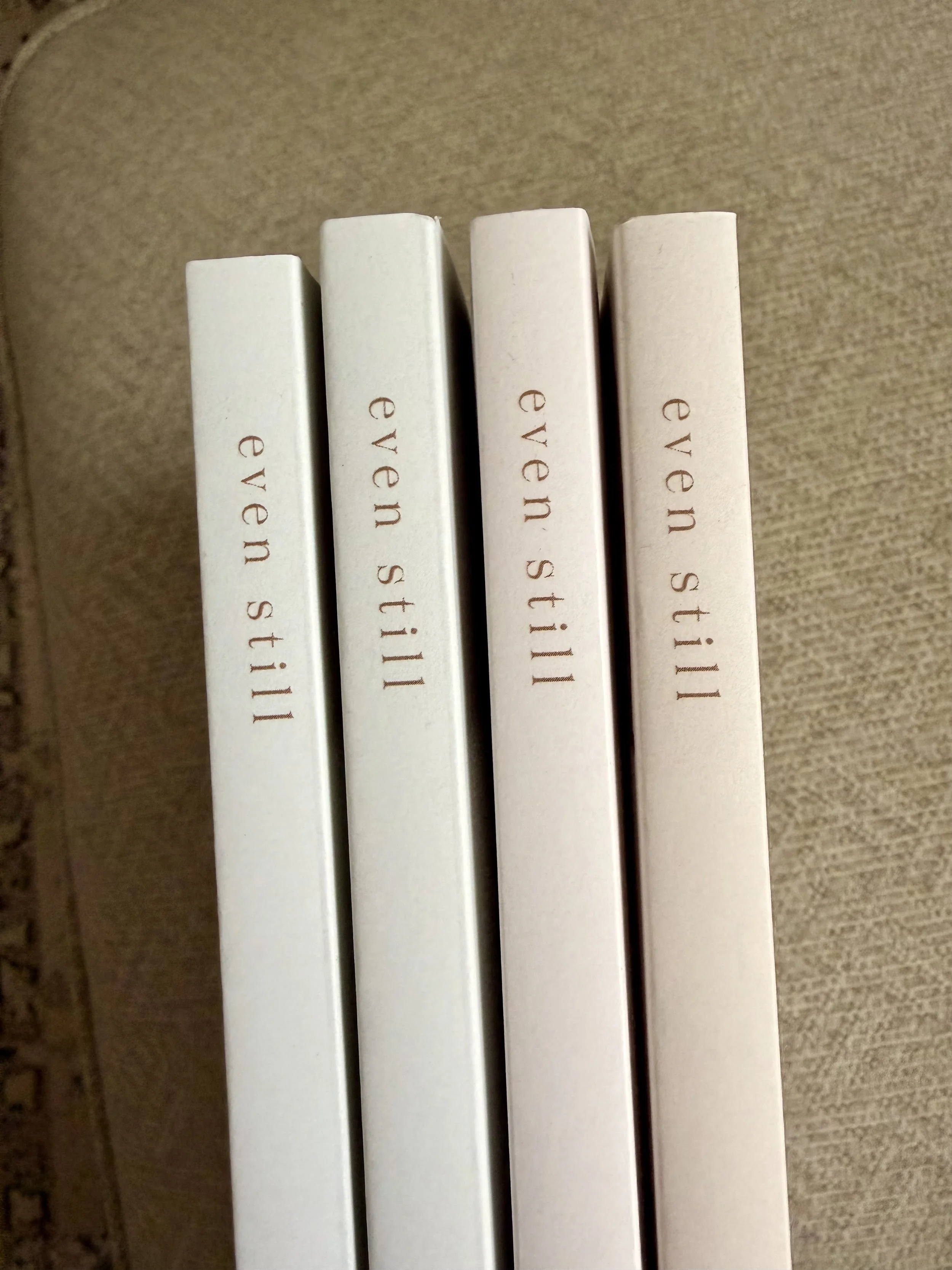 Close-up of four white books standing upright on a beige textured surface with the words "even-still" printed in simple font on the spines.