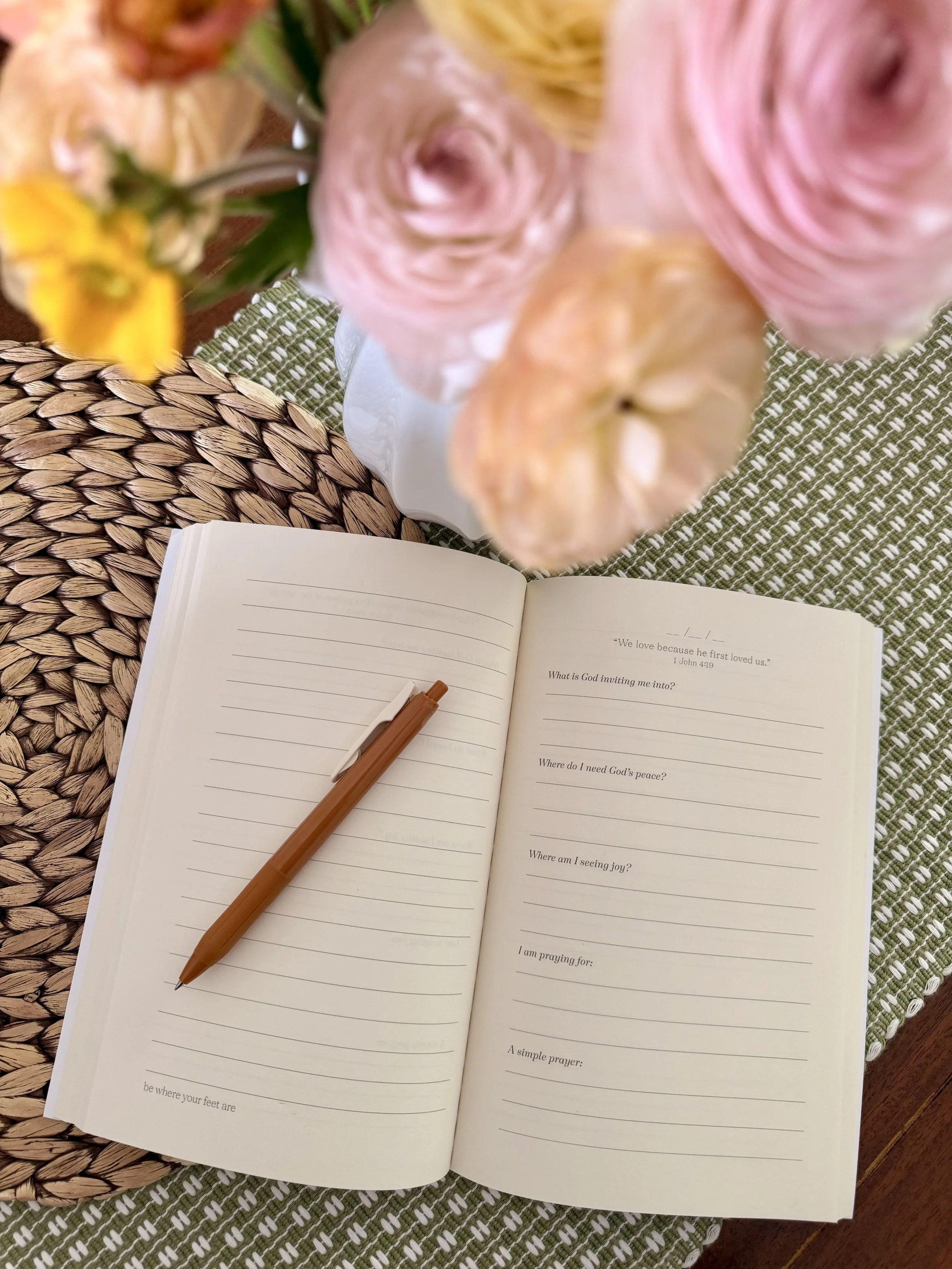 An open journal with lined pages and a brown pen resting on it, on a woven placemat and a green and white patterned cloth. There is a bouquet of flowers above the journal, and part of a decorative white plate is visible underneath the flowers.