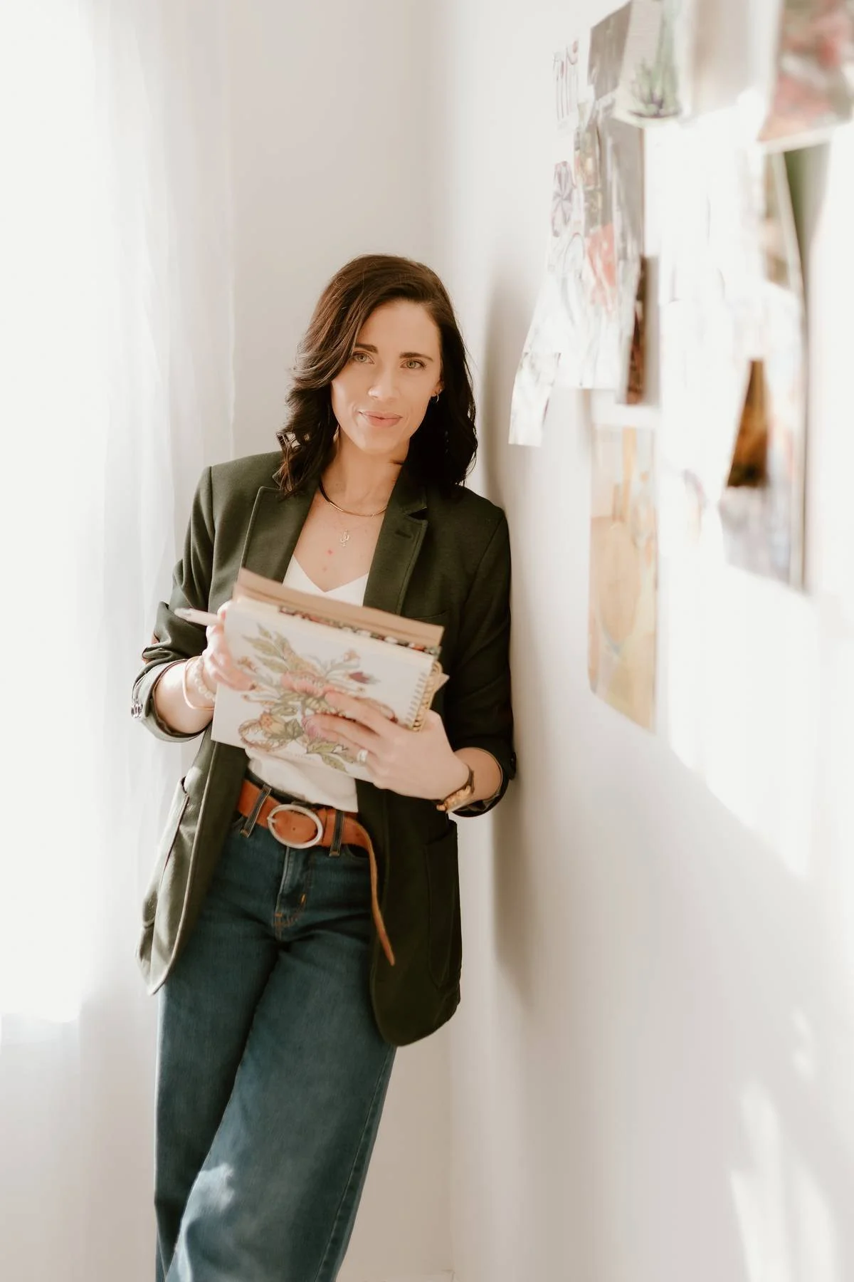 A woman with dark hair wearing a black blazer and jeans, holding a floral notebook, standing in a well-lit room with white walls, next to a wall displaying pictures and artwork.