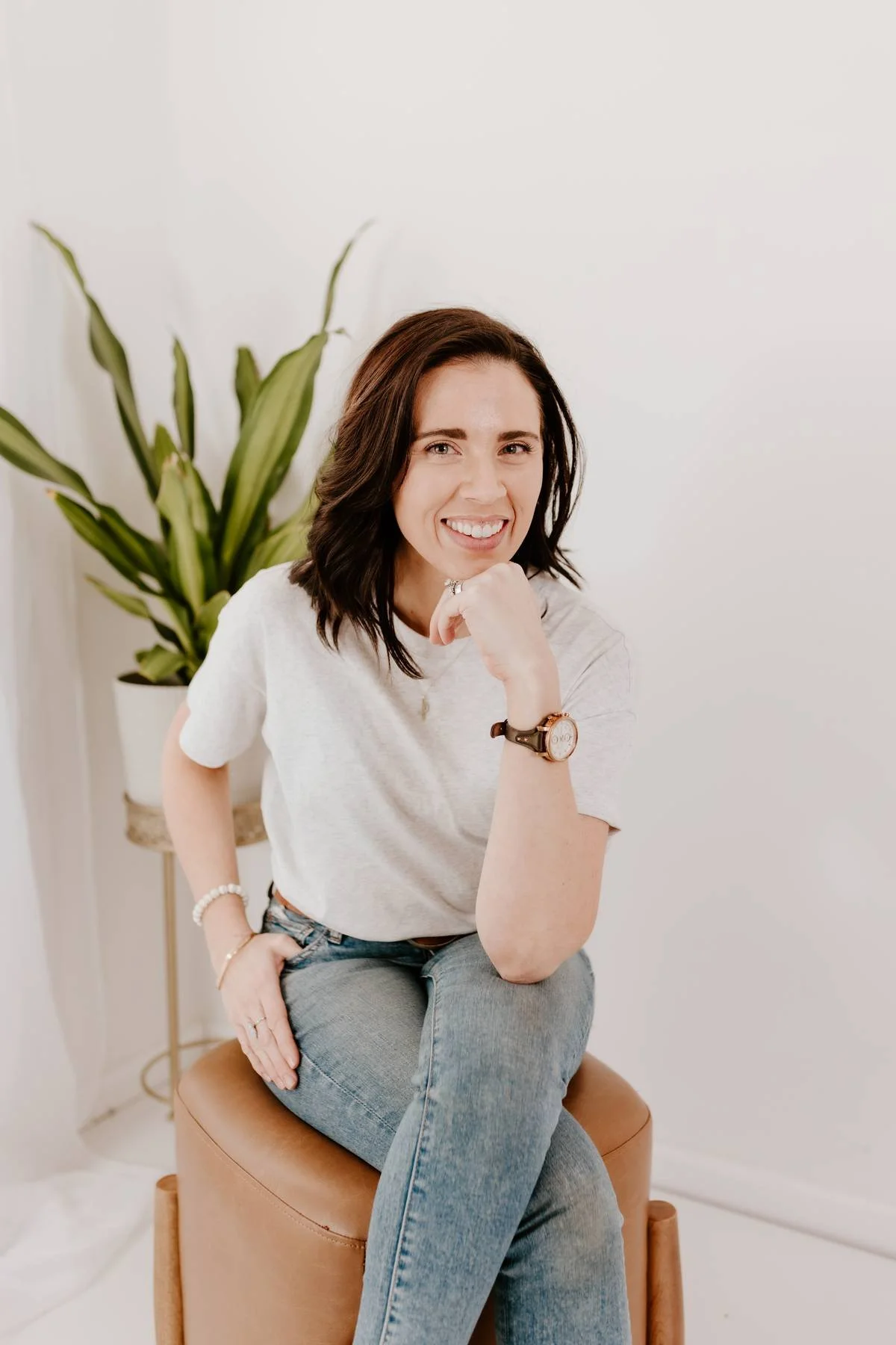 A woman with dark, shoulder-length hair smiling while sitting on a tan stool in front of a white wall and a tall plant with green leaves.