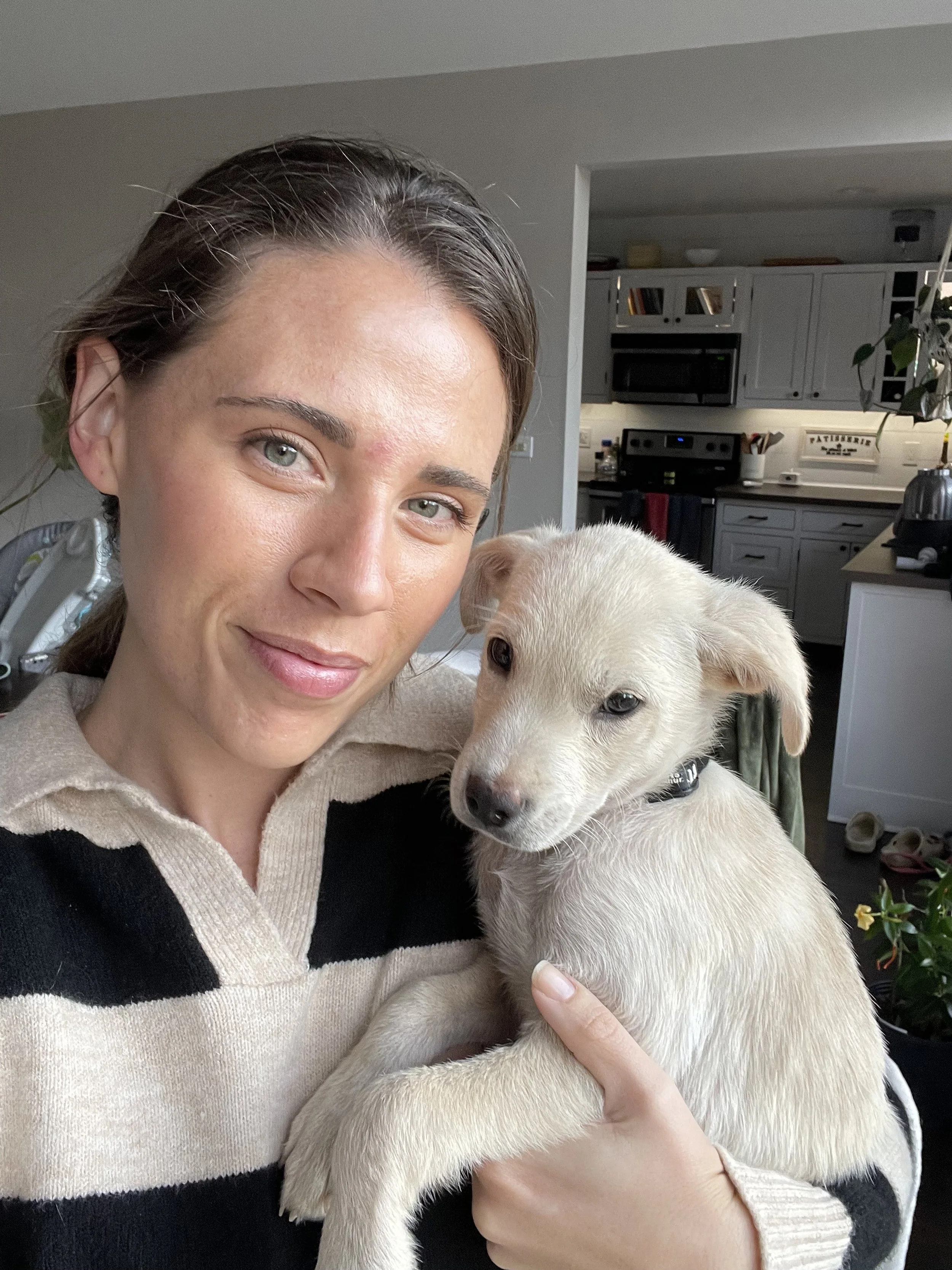 A woman with light skin and brown hair holding a light-colored puppy, standing inside a home with a kitchen in the background.