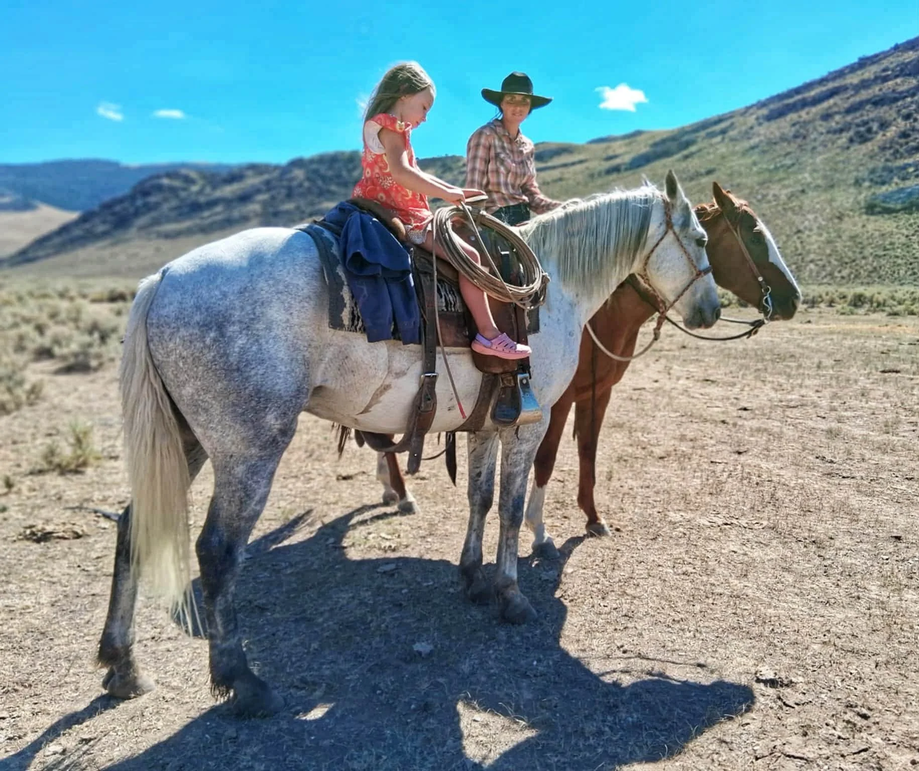A young girl riding a white horse while a woman, wearing a cowboy hat, rides a brown horse in a desert landscape with mountains in the background.