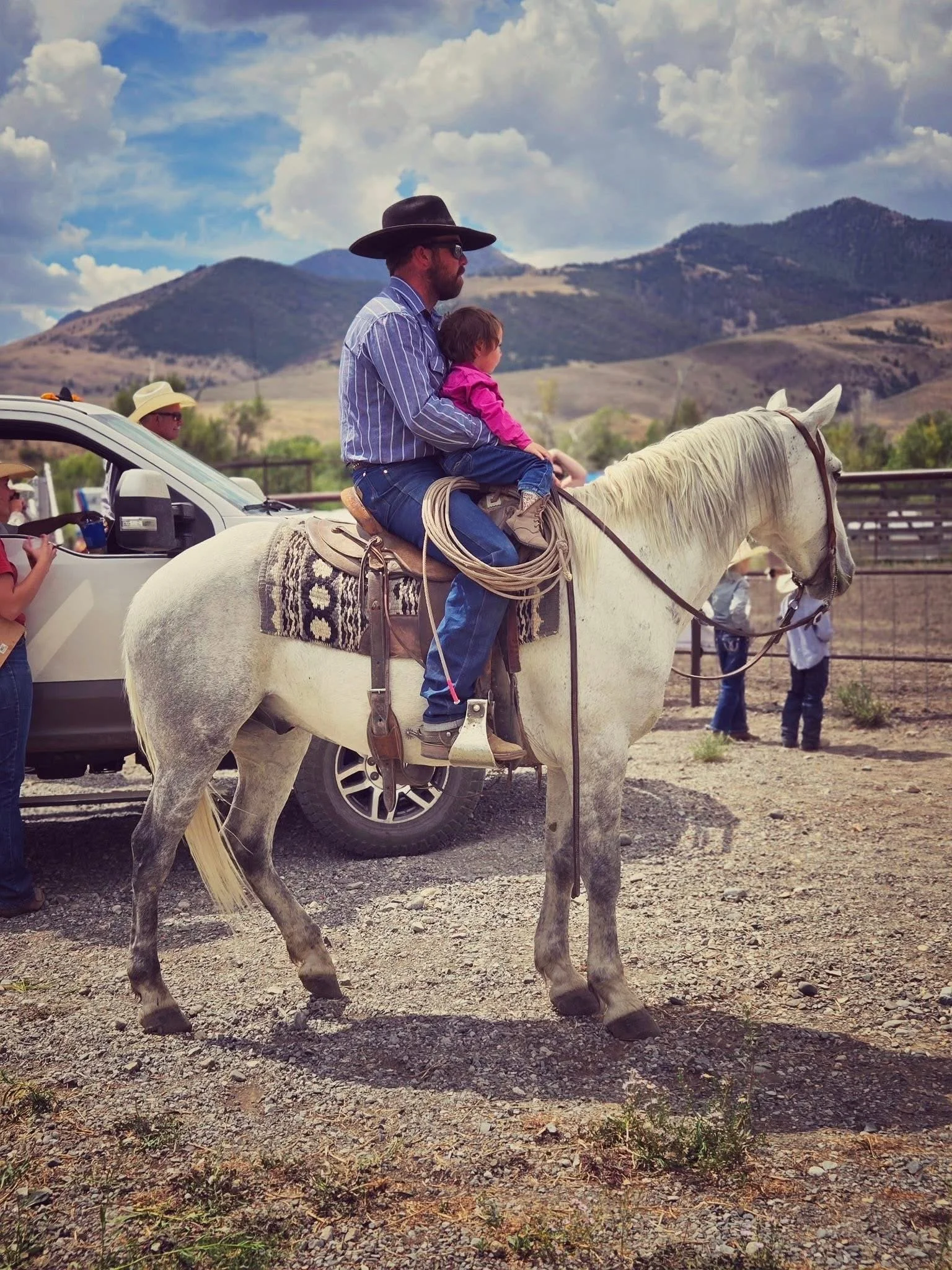 Man and young girl riding a white horse, with a man in a cowboy hat in the background and a rugged outdoor landscape with mountains.