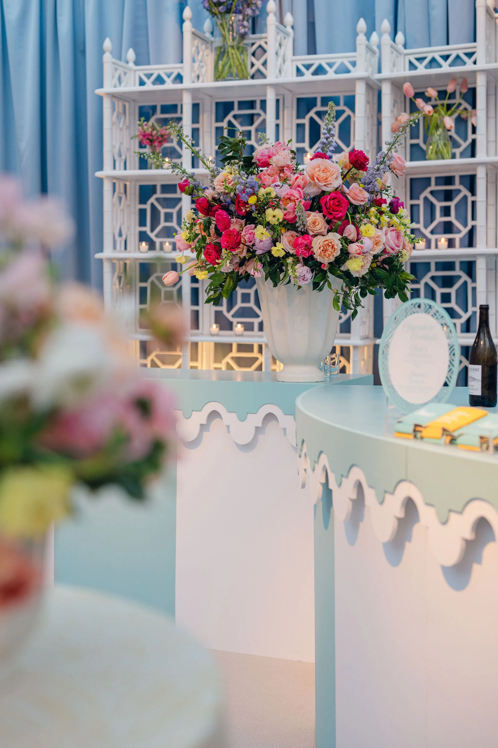 A large floral arrangement with pink, red, and cream roses, lavender, and greenery in a white vase on a pastel blue table, with a decorative white and blue tiered shelf with candles and vases in the background, set against blue drapes.