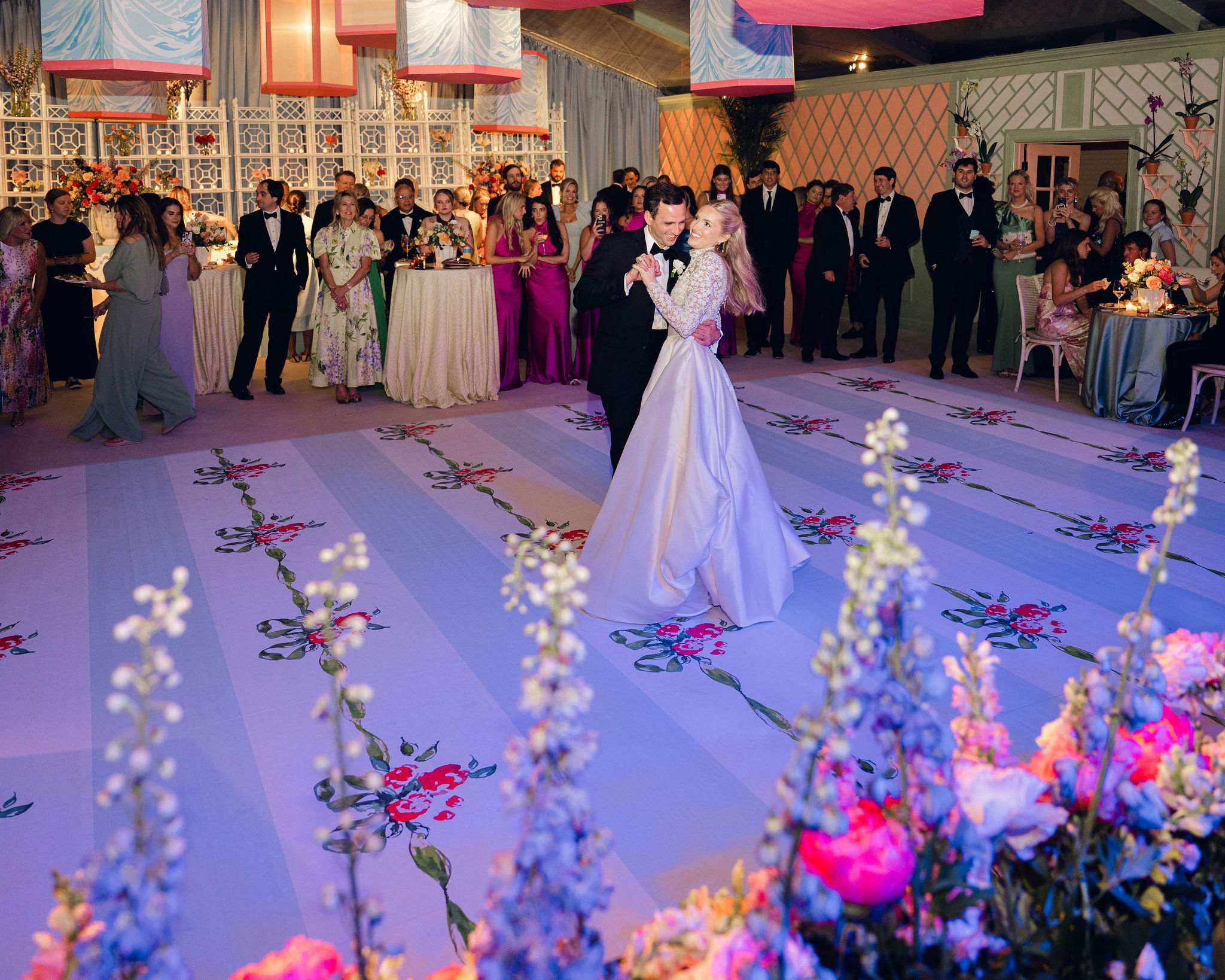 A bride and groom dancing at a wedding reception with guests watching and taking photos, decorated with floral arrangements and hanging lanterns.