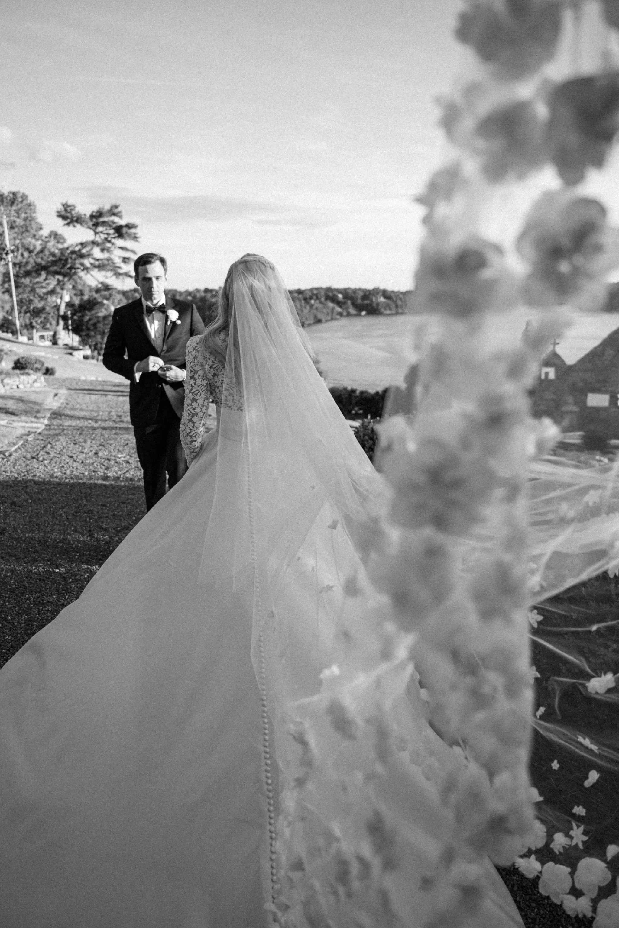 Black and white photo of a bride in a wedding dress and veil facing a groom in a tuxedo, outdoors with hills, trees, and a small church in the background, and flower petals scattered on the ground.