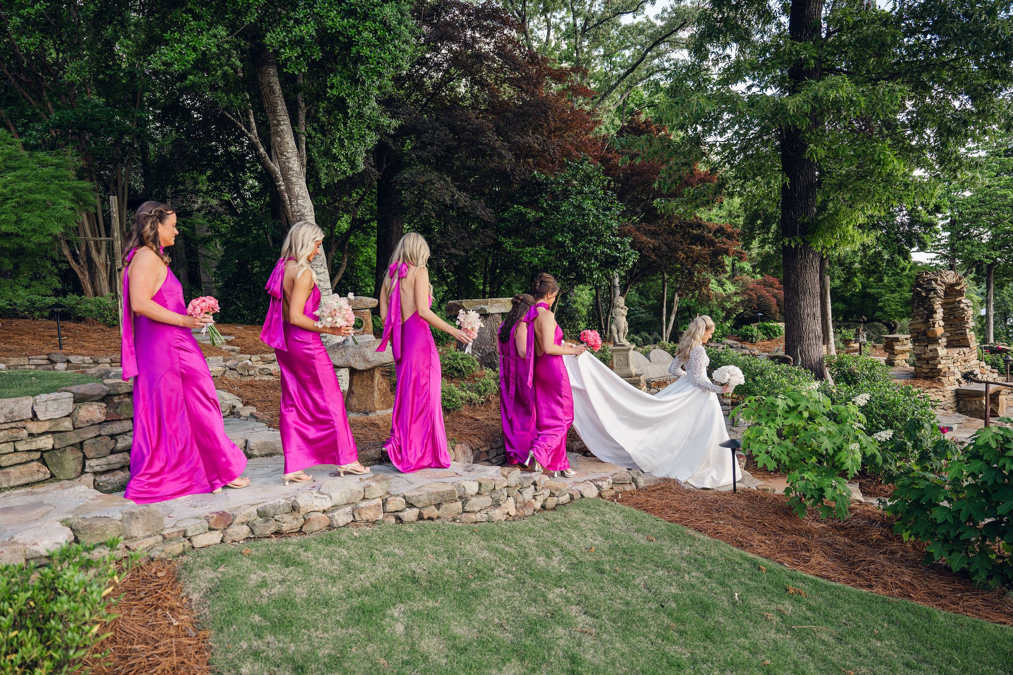 A group of women dressed in bright fuchsia dresses and a woman in a white wedding gown walking in a garden setting, holding bouquets of flowers. The scene is outdoors with lush green trees and foliage.