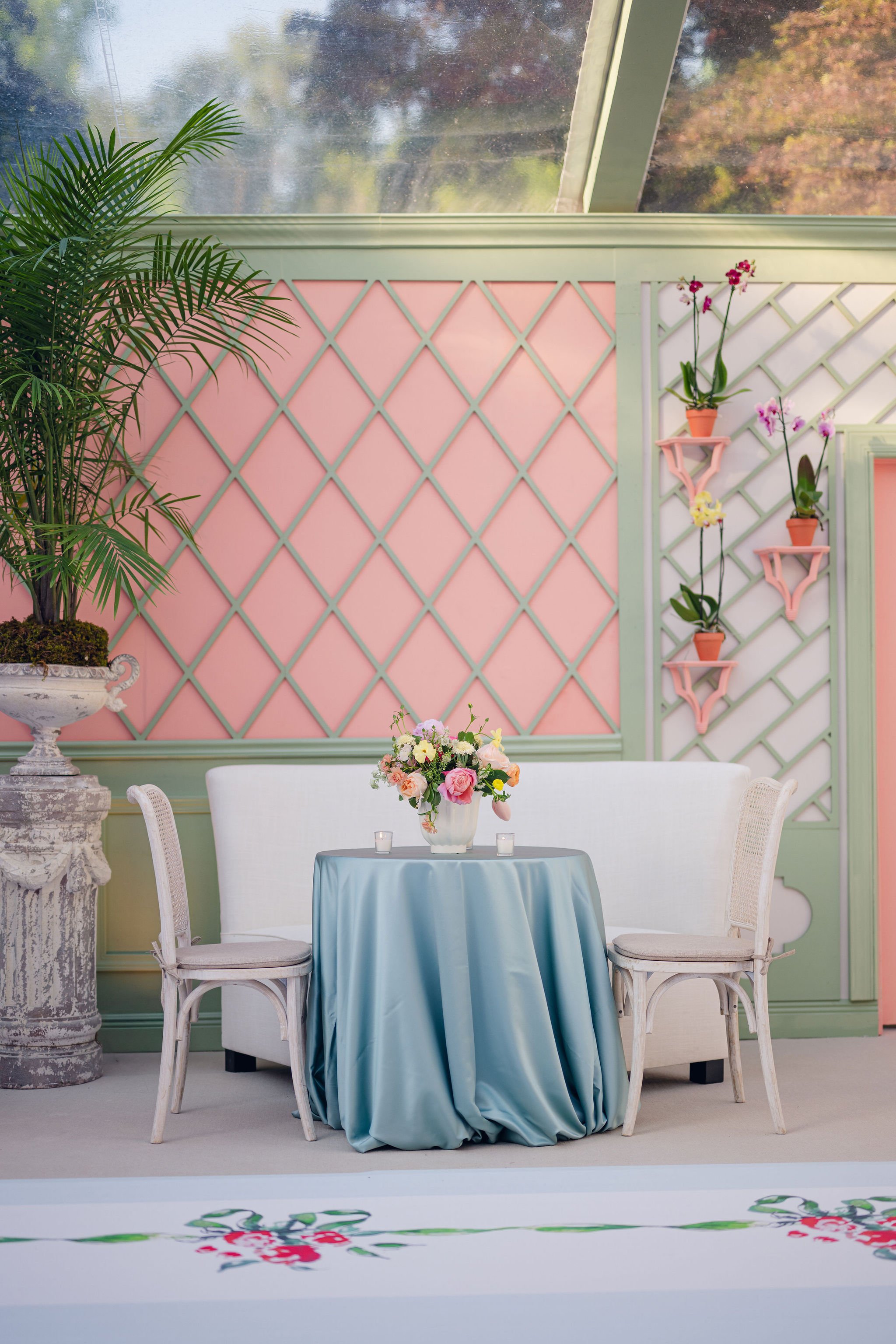 A round table covered with a light blue tablecloth, with a floral centerpiece. Four white chairs surround the table. Behind, a pink and mint green lattice wall decorates the space, with potted orchids hanging on pink shelves. A large green potted pla