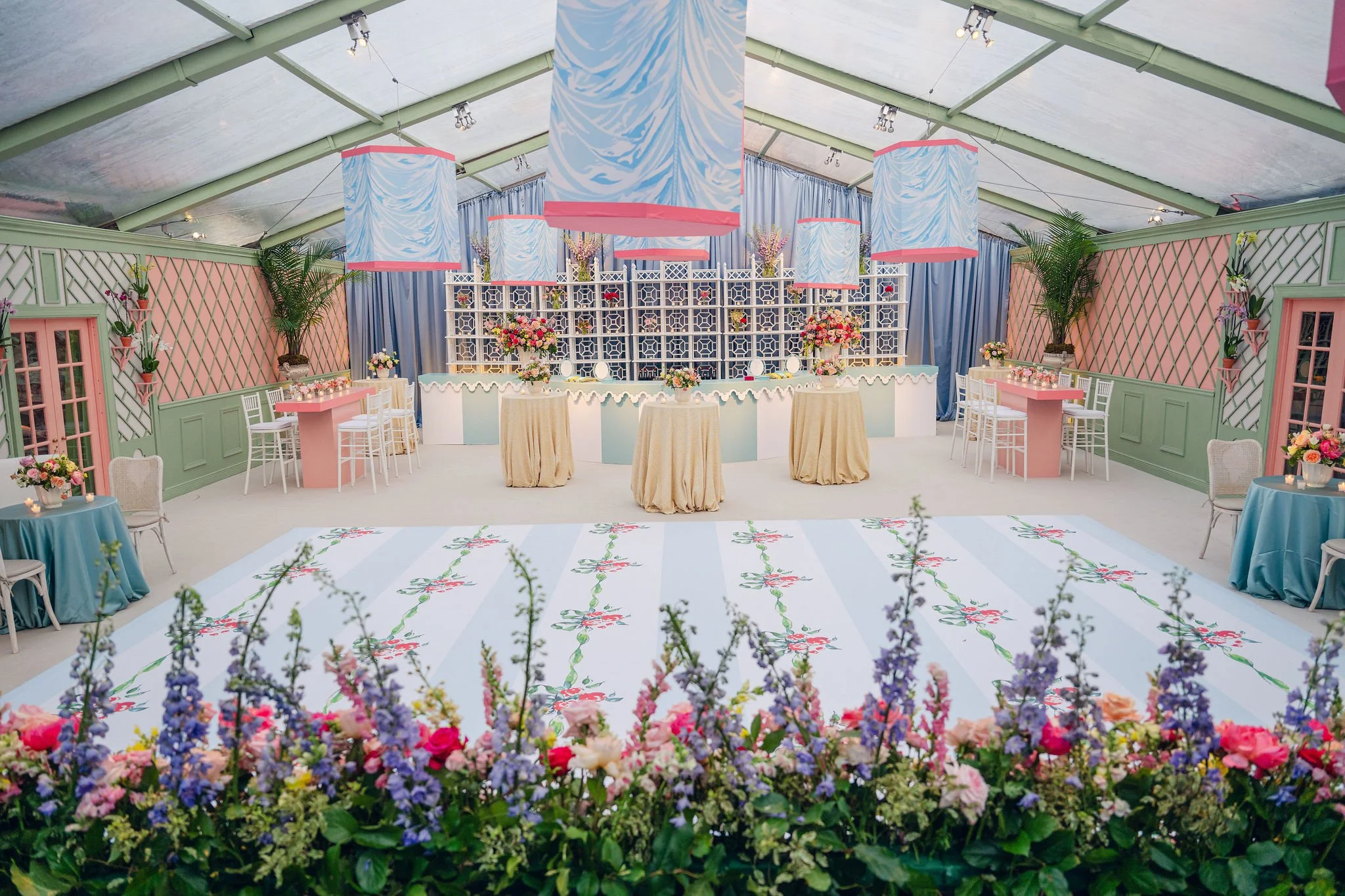 A colorful indoor venue decorated for a celebration. The ceiling is covered with a tent-like structure, and the room features pastel green walls with pink accents. There are large hanging lamps with blue and pink swirled designs, and various tables d
