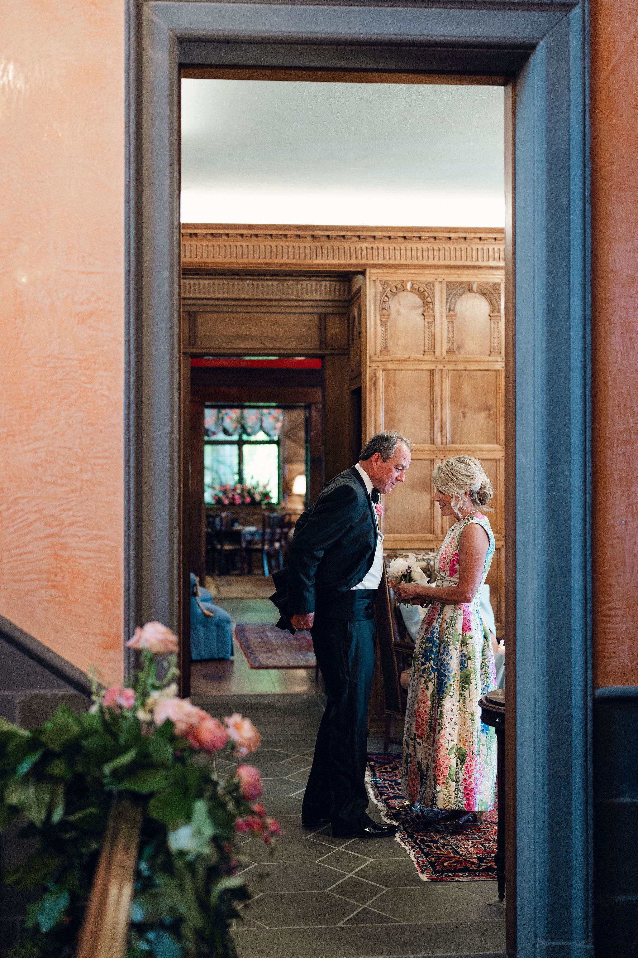 A couple, dressed formally, stands together in an elegant, wood-paneled room with a patterned carpet, sharing an intimate moment, possibly during a wedding or special occasion, seen through a doorway with pink walls and flowering plant in the foregro