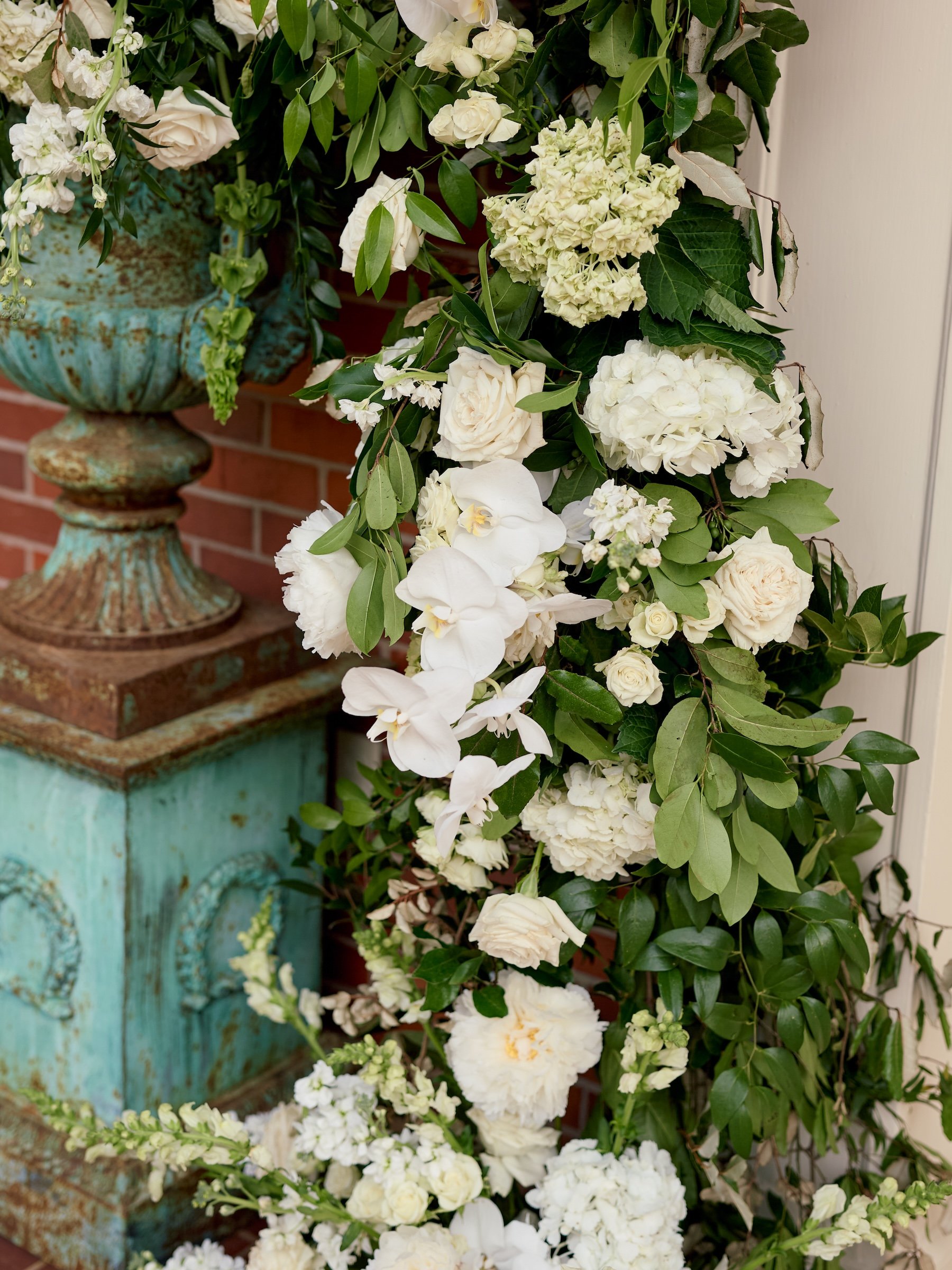 Various white flowers, including roses and orchids, arranged with green leaves around a rustic, turquoise urn with a weathered finish.