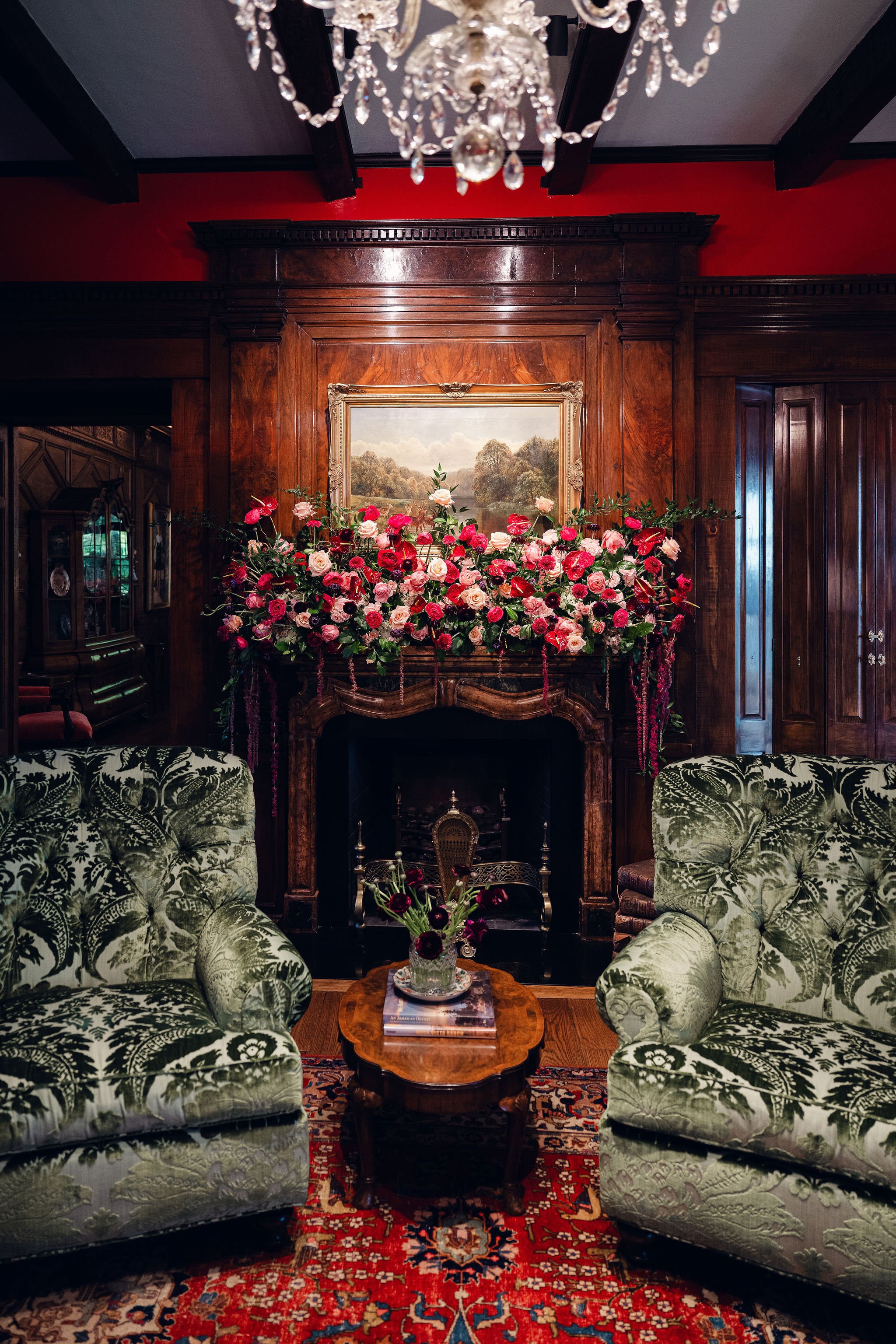 A vintage living room with a wooden fireplace mantel decorated with a large floral arrangement of pink and red roses and greenery. Above the mantel, there is a landscape painting. On top of the mantel, there is a marble clock. Two upholstered armchai