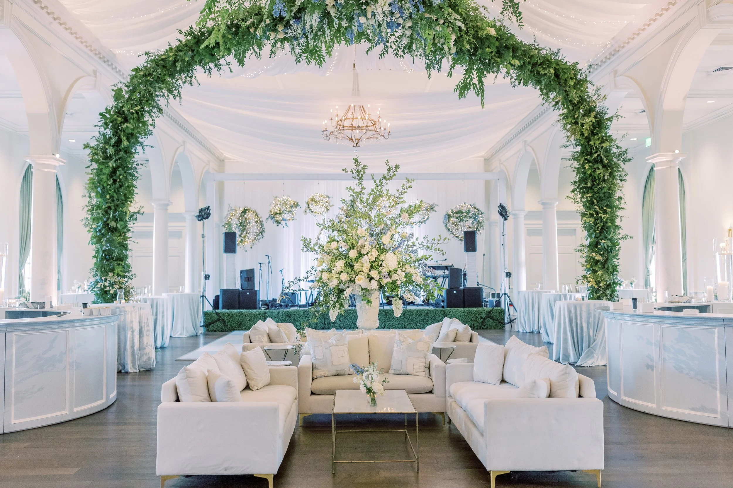 Elegant wedding or event reception hall decorated with a large floral arrangement at the center, greenery arch overhead, chandeliers, and a stage with floral wreaths and sound equipment.