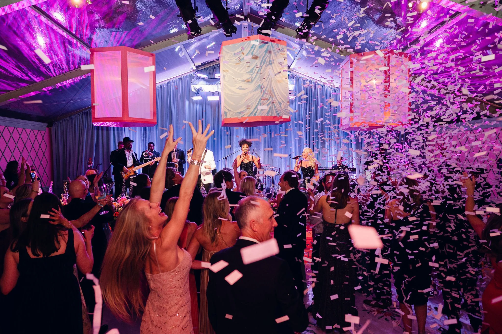 People at an event celebrating, with confetti falling from the ceiling, a band performing on stage, and colorful lighting.