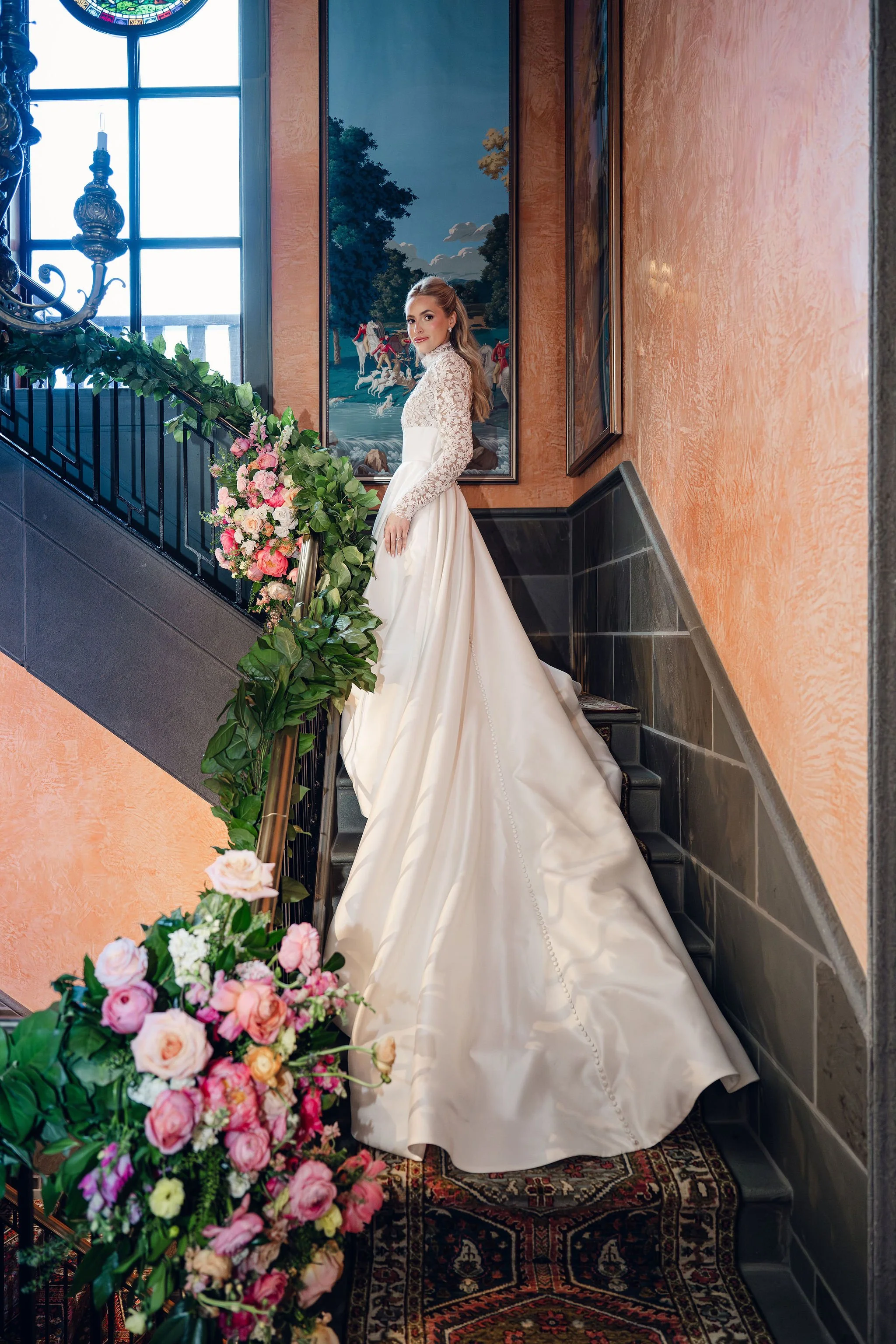 A woman in a white wedding dress standing on stairs decorated with pink and white flowers, inside a building with orange textured walls, black tiled accents, and large windows, with paintings on the wall behind her.