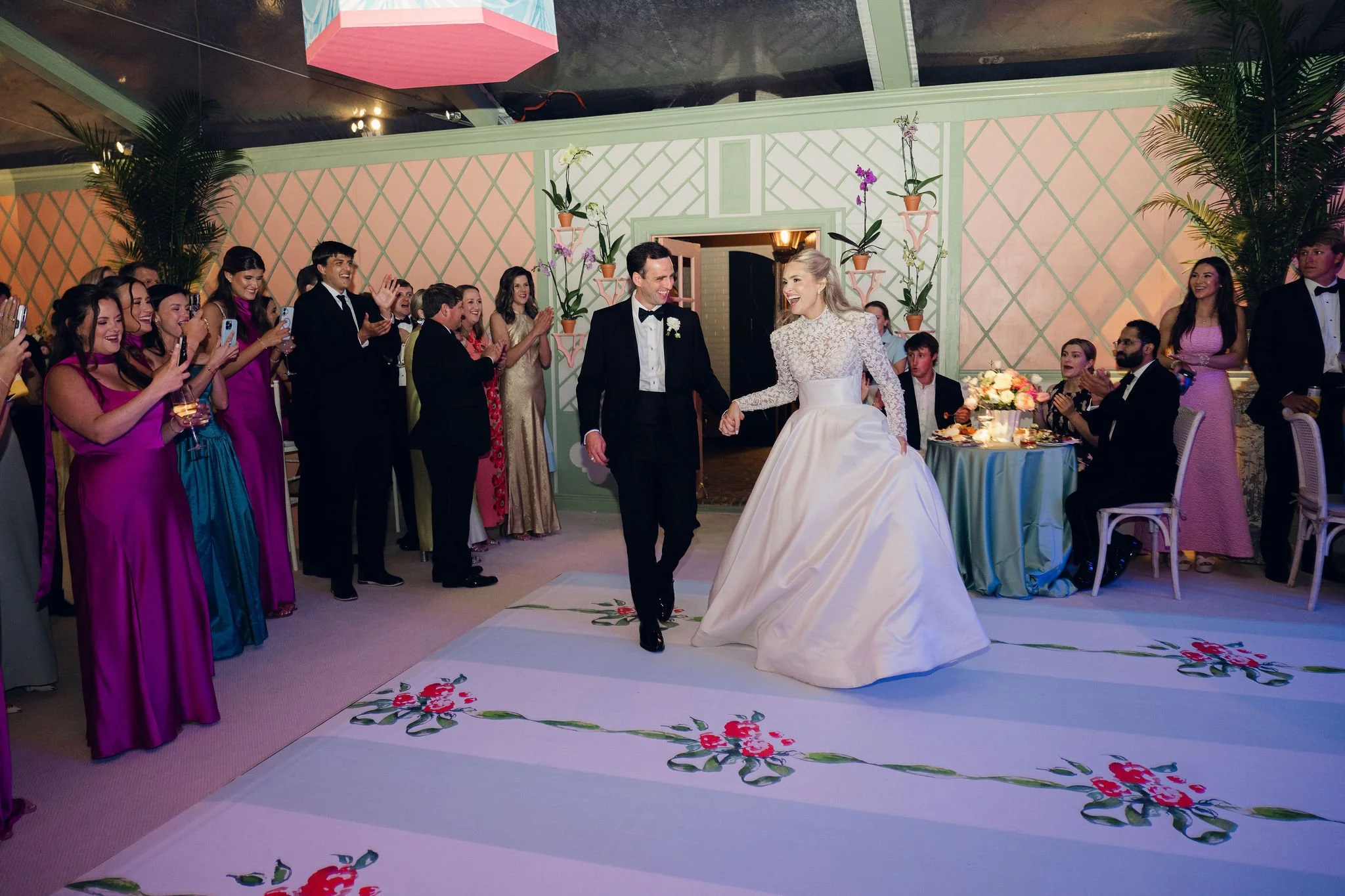 A wedding reception with a bride and groom dancing in front of guests taking photos and clapping, decorated with plants and floral arrangements.