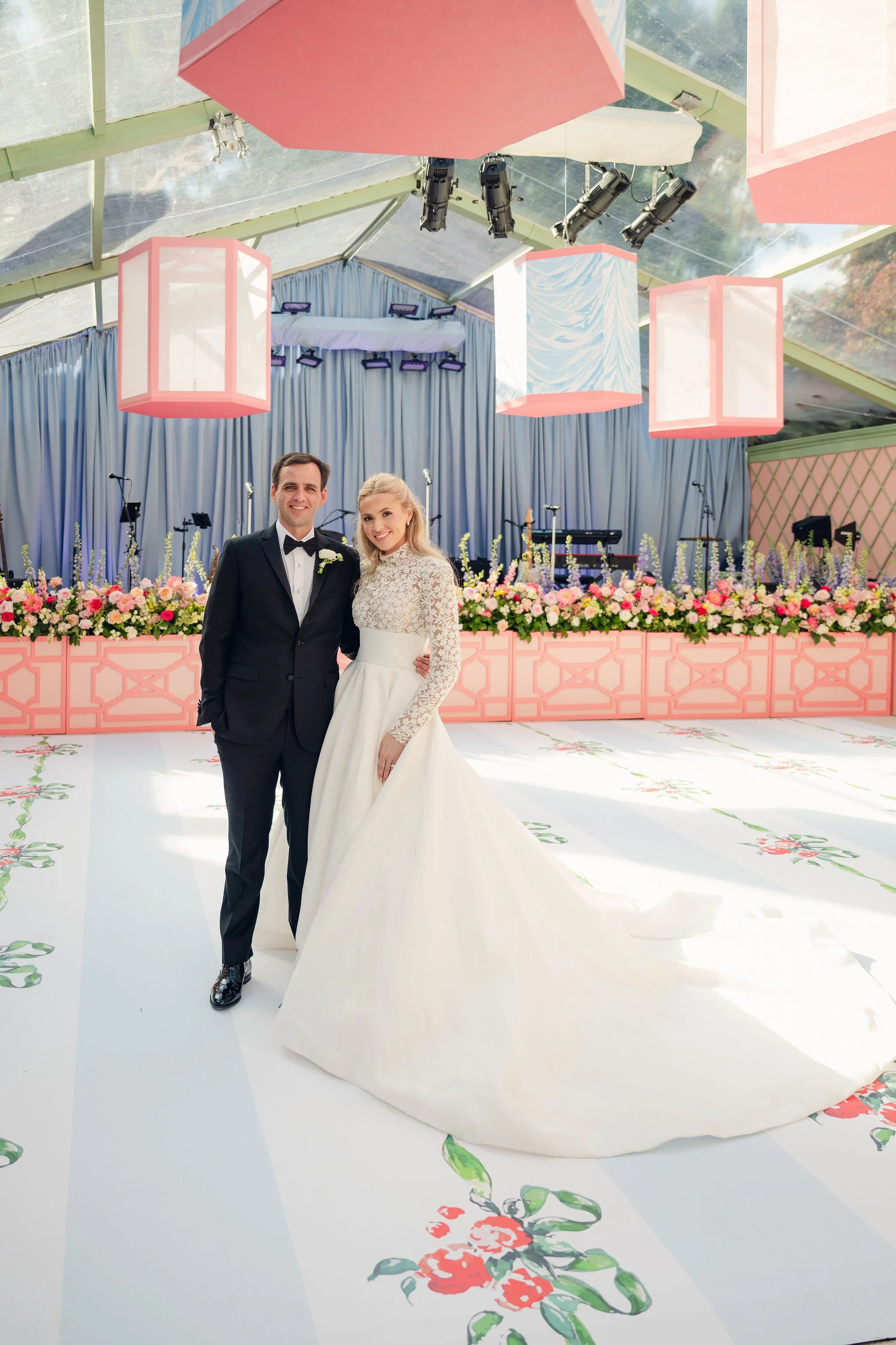 A bride and groom standing together at a wedding reception, smiling. The bride is wearing a white lace wedding gown and the groom is in a black tuxedo. Behind them is a decorated stage with pink and purple flowers and a blue curtain. The ceiling has 