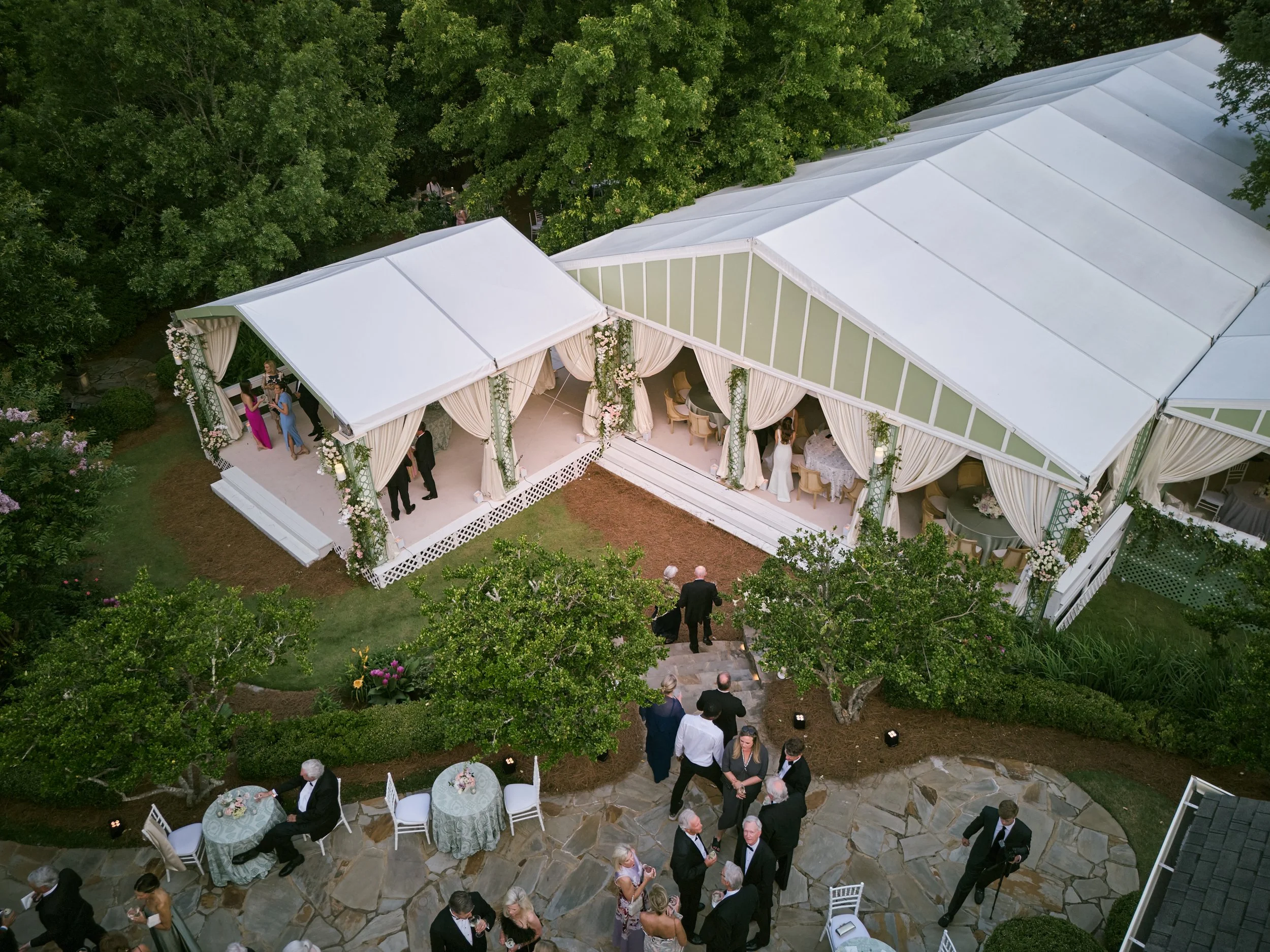 Aerial view of a wedding reception outdoors with a white tent, surrounded by trees, and people mingling and seated at tables.