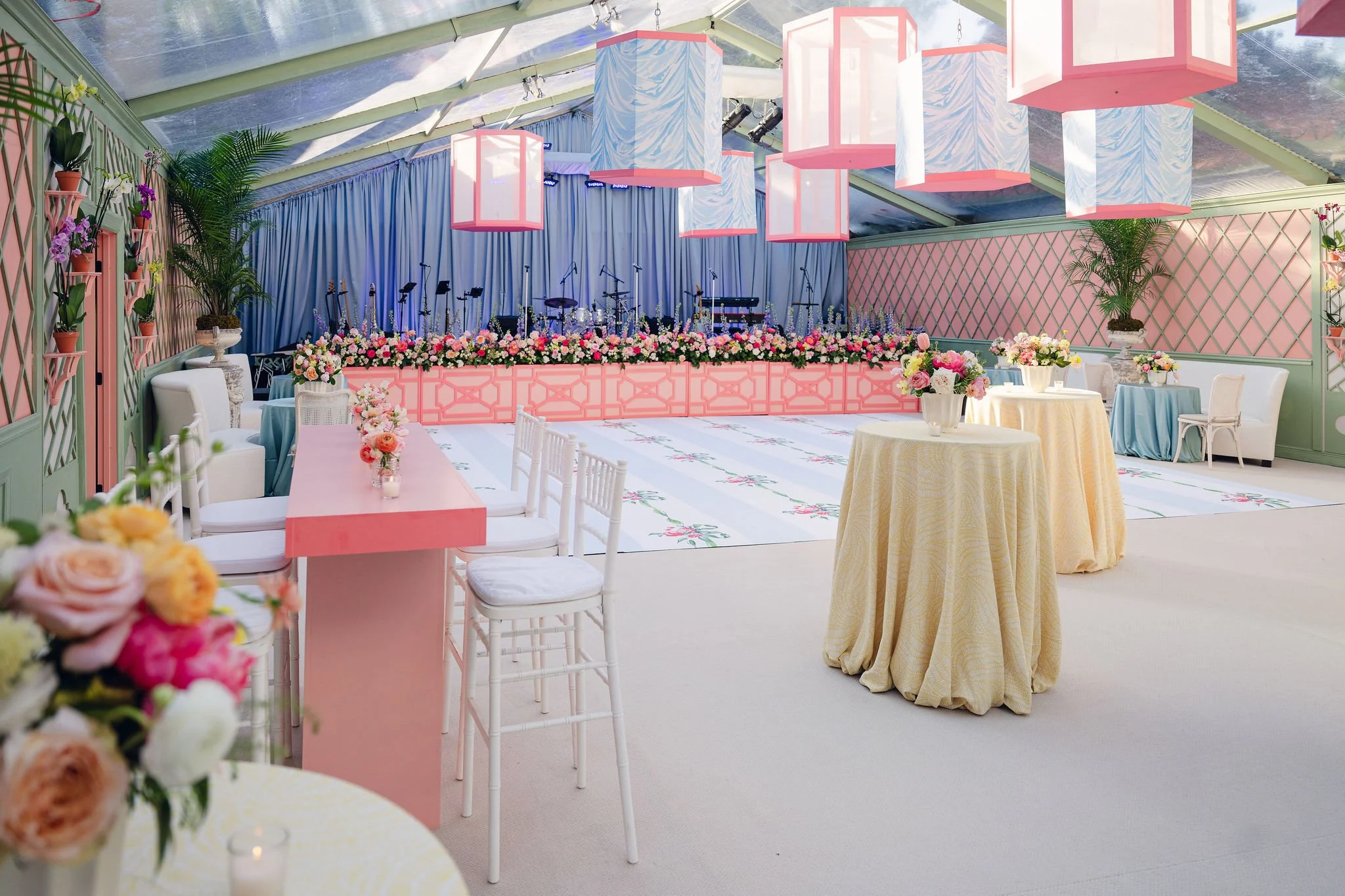 Decorated event space with pastel pink and blue color scheme, floral arrangements, round tables with tablecloths, and a stage with musical instruments under hanging geometric lanterns inside a clear tent.