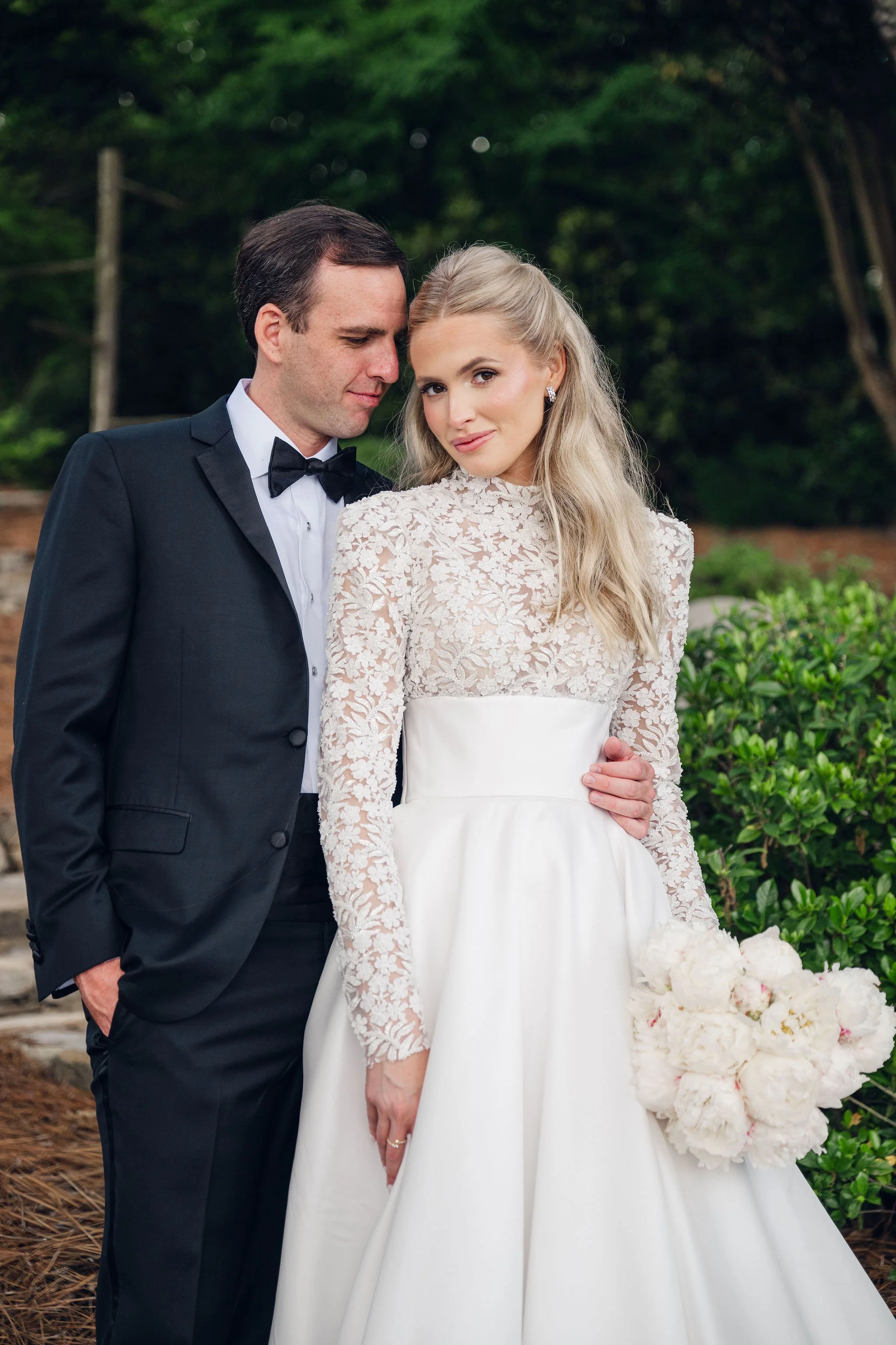 A bride and groom at their wedding, standing outdoors with greenery in the background. The groom is wearing a black tuxedo with a bow tie, and the bride is in an elegant white lace wedding dress, holding a bouquet of white flowers.