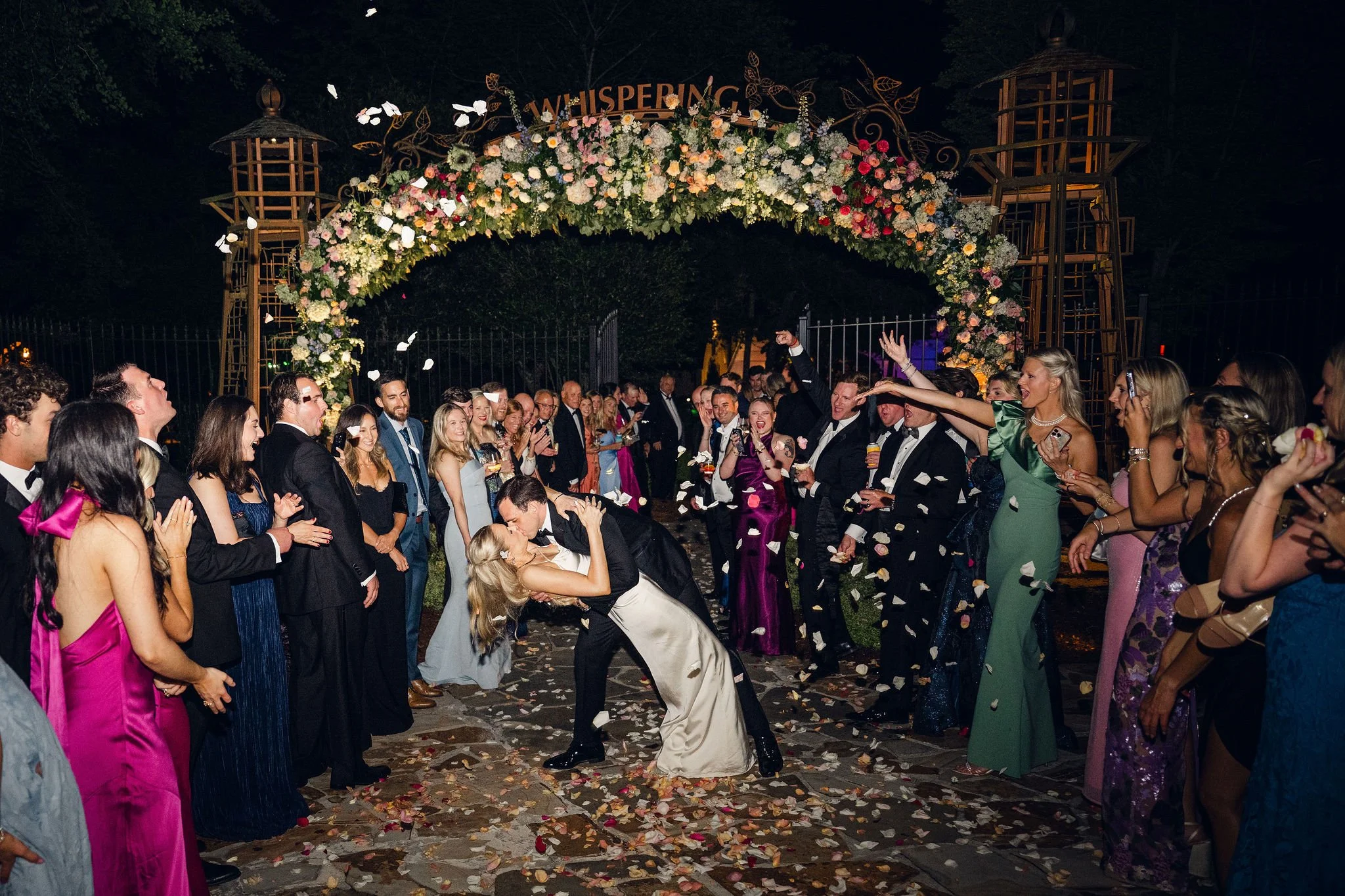 Nighttime wedding celebration with guests throwing flower petals, smiling, dressed in formal attire, under a floral archway labeled 'Whispering' with lanterns on either side.