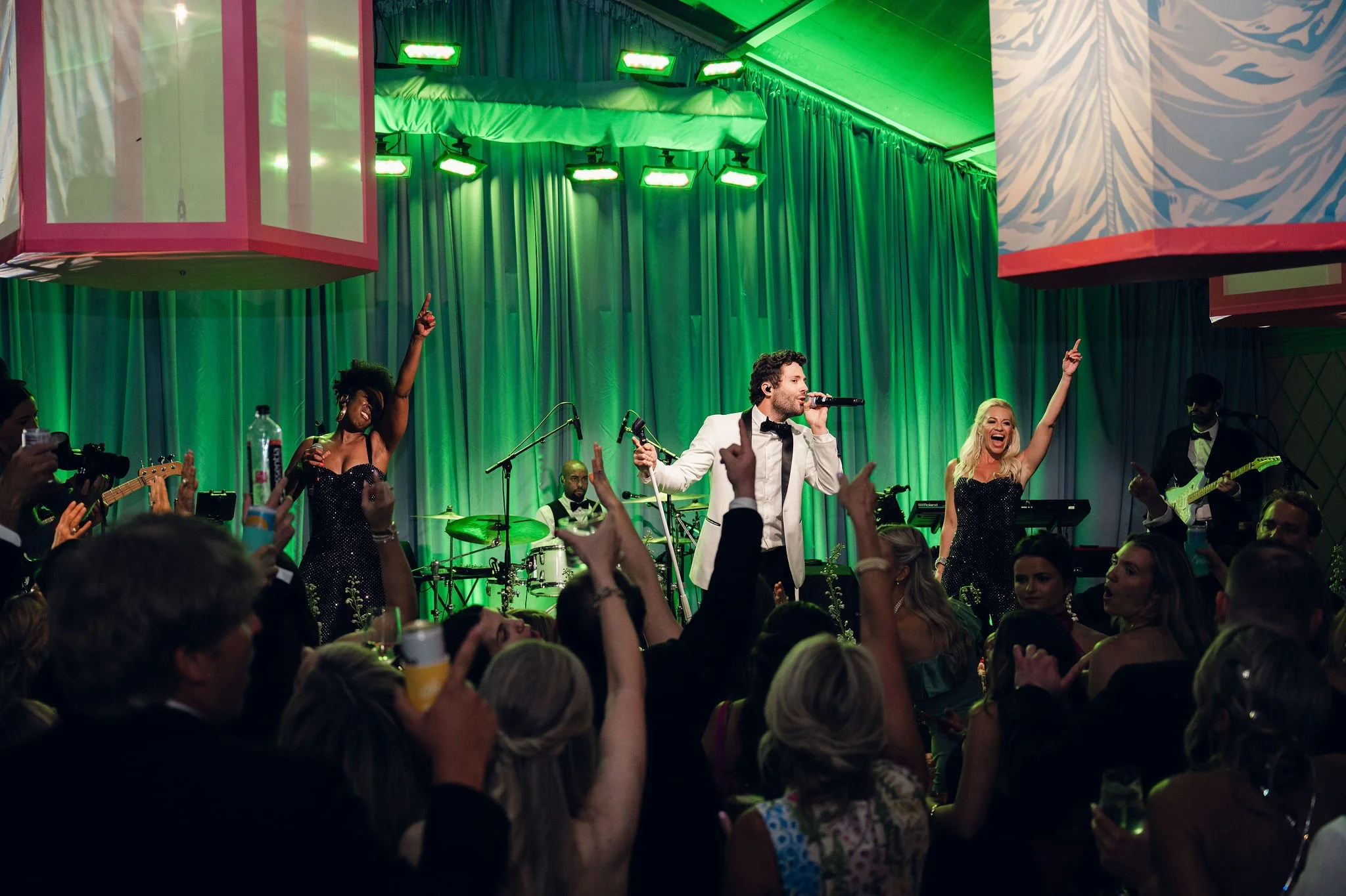 Performance on stage at a lively event with musicians, singers, and audience members raising their hands, illuminated by green stage lighting.