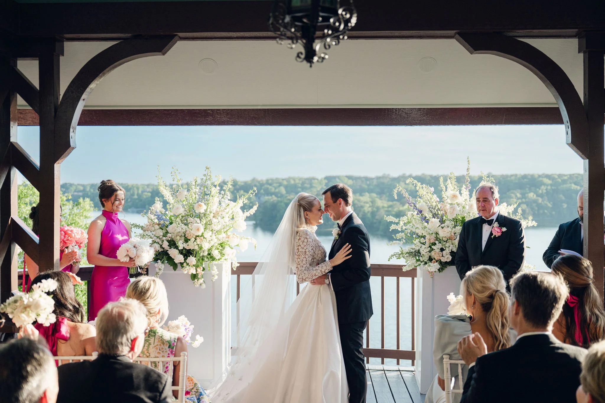 An outdoor wedding ceremony with a bride and groom exchanging vows under a wooden arch decorated with large white flowers, with guests seated facing them, overlooking a scenic lake and green landscape in the background.