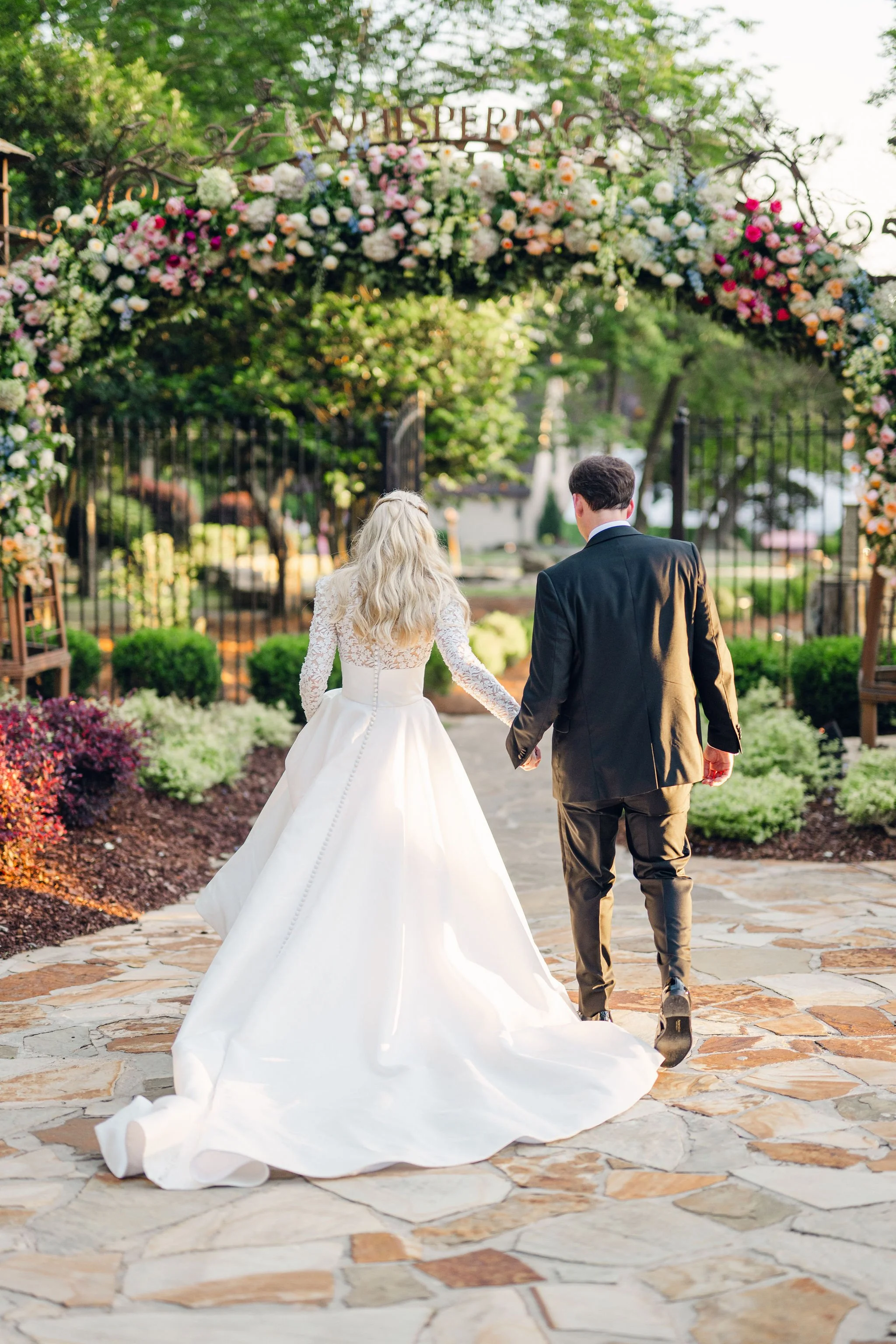 A bride and groom walking hand in hand under a floral arch at sunset.