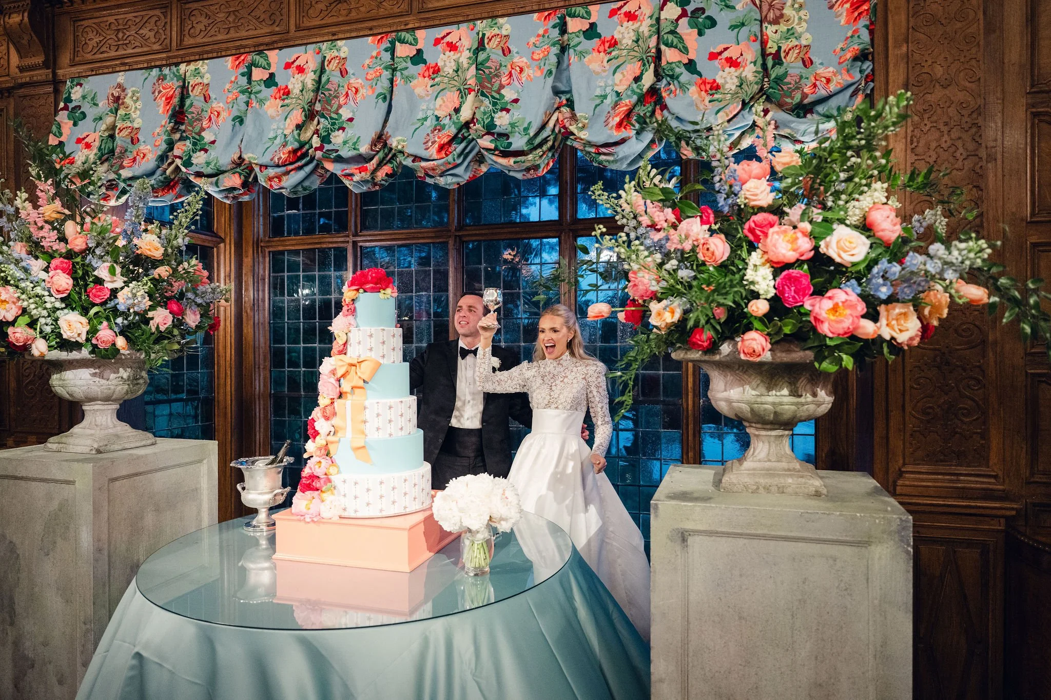 A bride and groom cutting a wedding cake at a reception hall decorated with large floral arrangements and floral curtains.