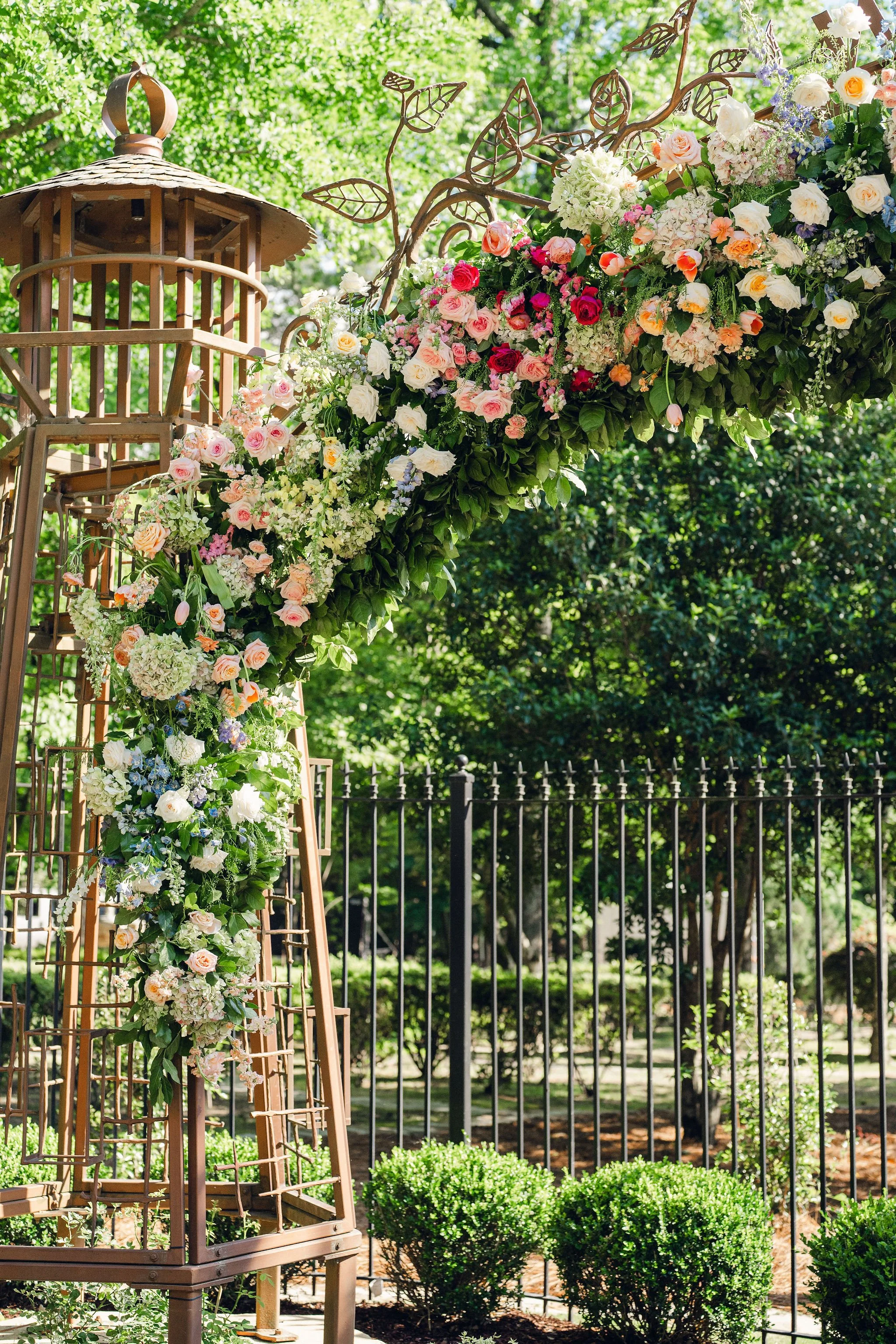 A garden arch decorated with pastel and white roses, hydrangeas, and greenery, with a wooden lantern on the left side, set in a lush green outdoor setting.