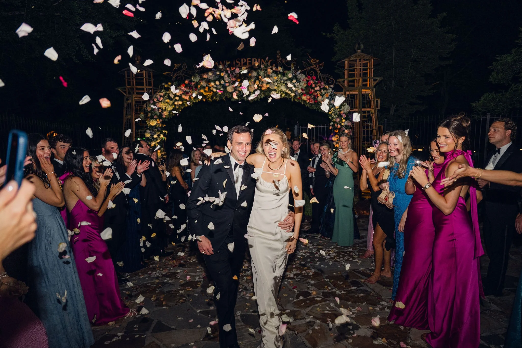 Couple in wedding attire smiling and celebrating with guests at night, flower petals falling, under a decorated floral arch.