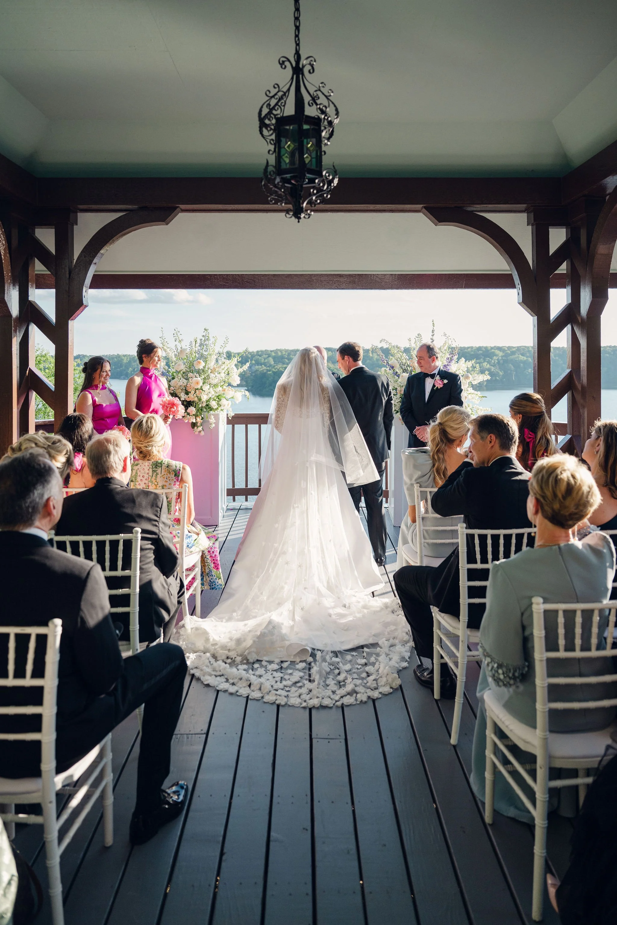 A wedding ceremony taking place on a porch overlooking a lake. The bride and groom stand facing each other, surrounded by bridesmaids and groomsmen, with guests seated nearby.