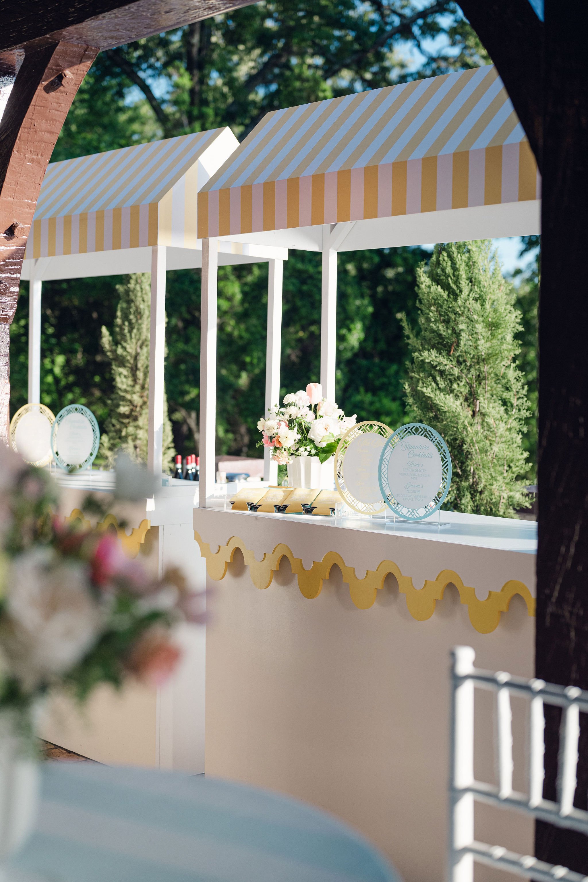 A decorated outdoor event setup with white and yellow striped canopies, tables with floral centerpieces, and signs in elegant frames, surrounded by green trees.