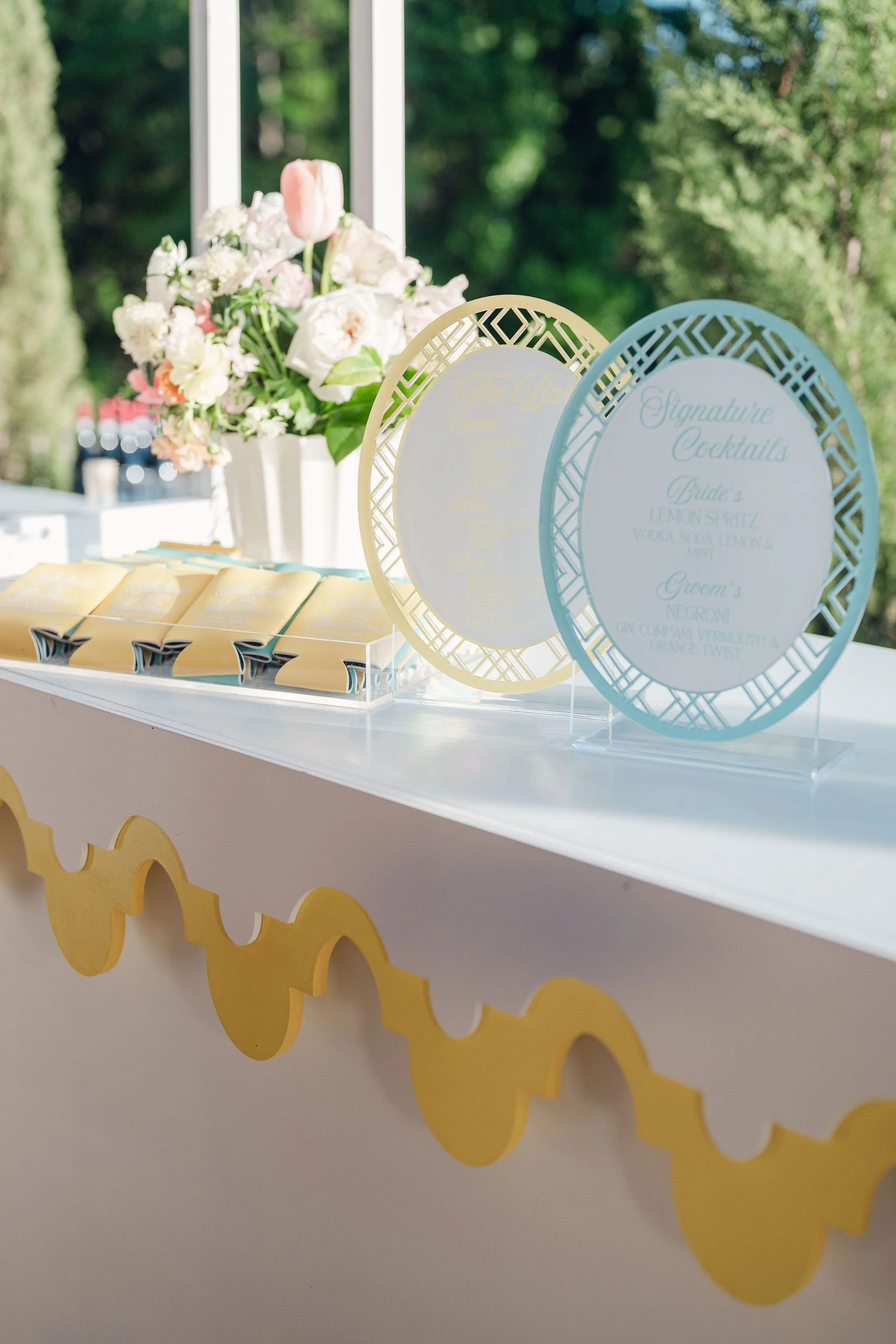 A white table decorated with a bouquet of various pink and white flowers in a white vase and a menu with blue and yellow frames, set outdoors against green trees and a clear sky.