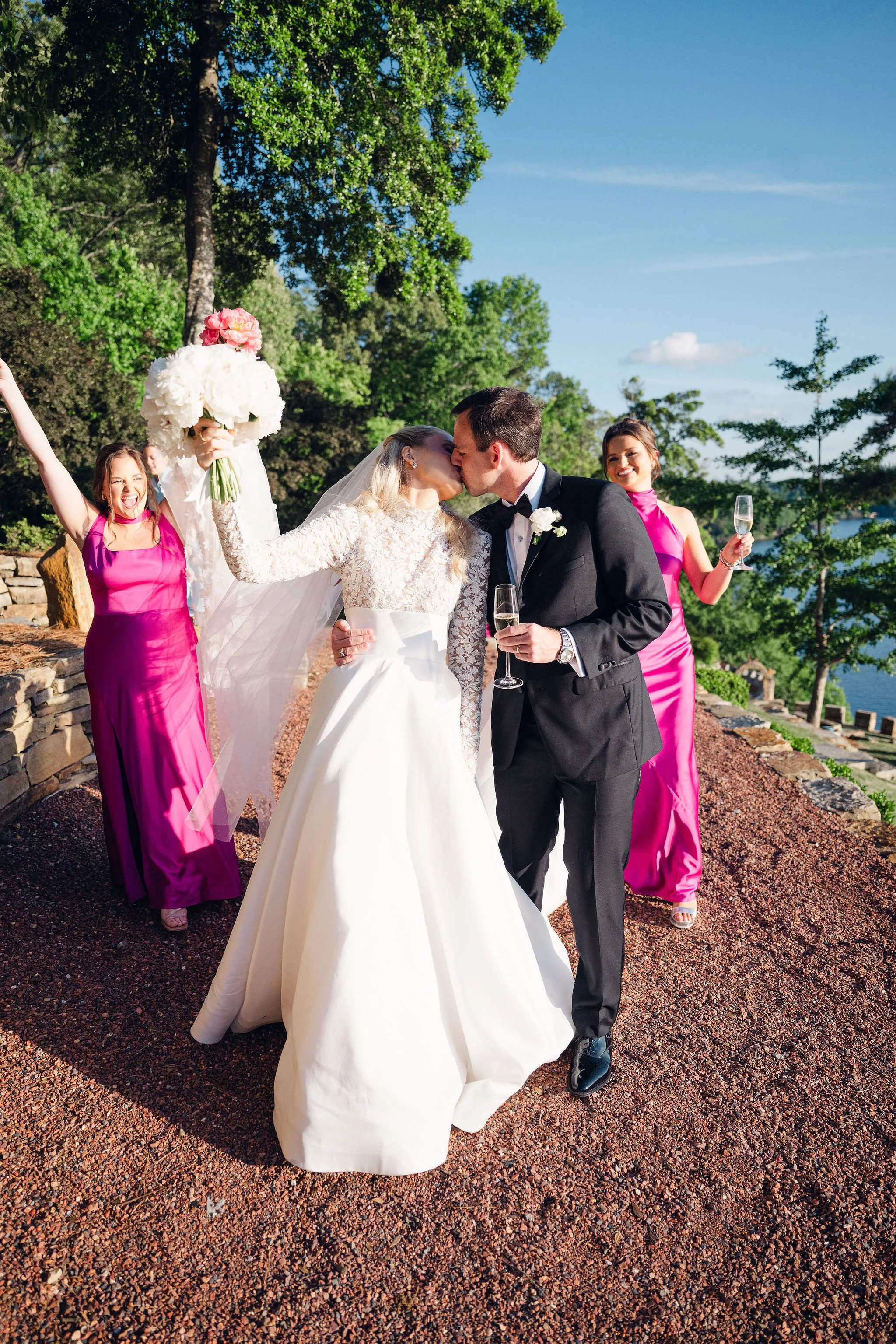 A bride and groom kiss at their wedding outside on a sunny day, with women in pink dresses celebrating nearby, holding glasses of champagne and smiling.