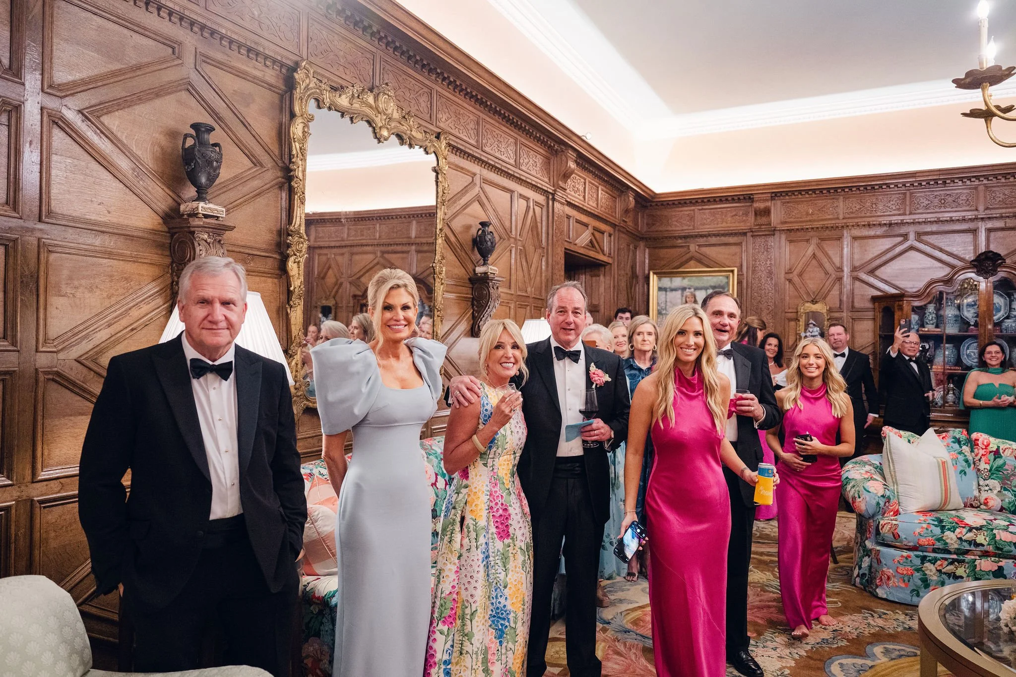 A group of people dressed in formal attire at a party or celebration in a wood-paneled room with ornate decorations.