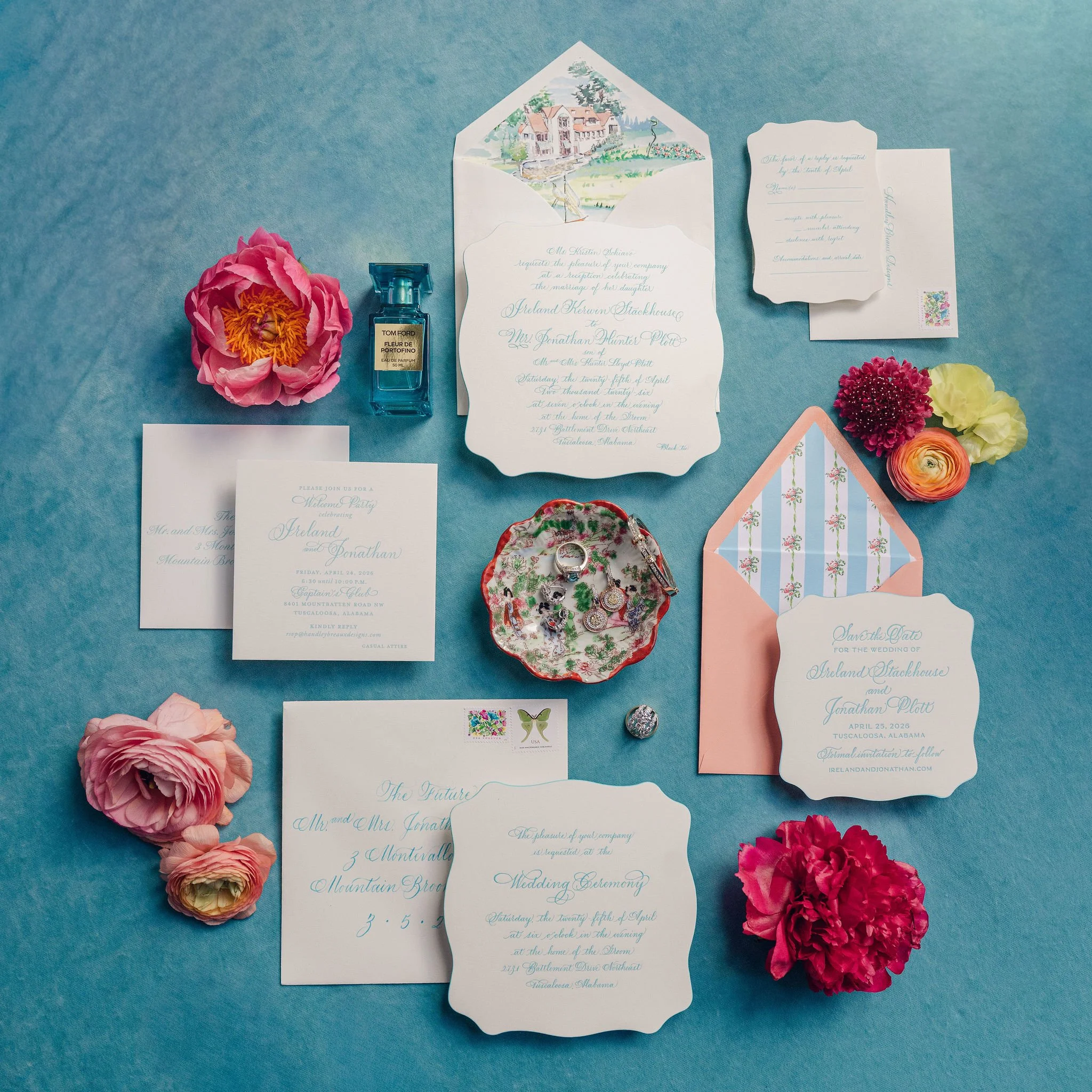 Wedding invitation suite with floral accents and a watercolor illustration of a house on the back of the main invitation, with pink and red flowers, a bottle of perfume, a compact mirror, and a pink and blue striped paper on a teal background.
