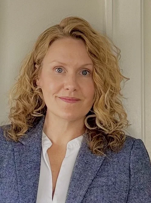 A woman with curly blonde hair and blue eyes wearing a white blouse and a gray blazer, smiling slightly, standing indoors in front of a white door.