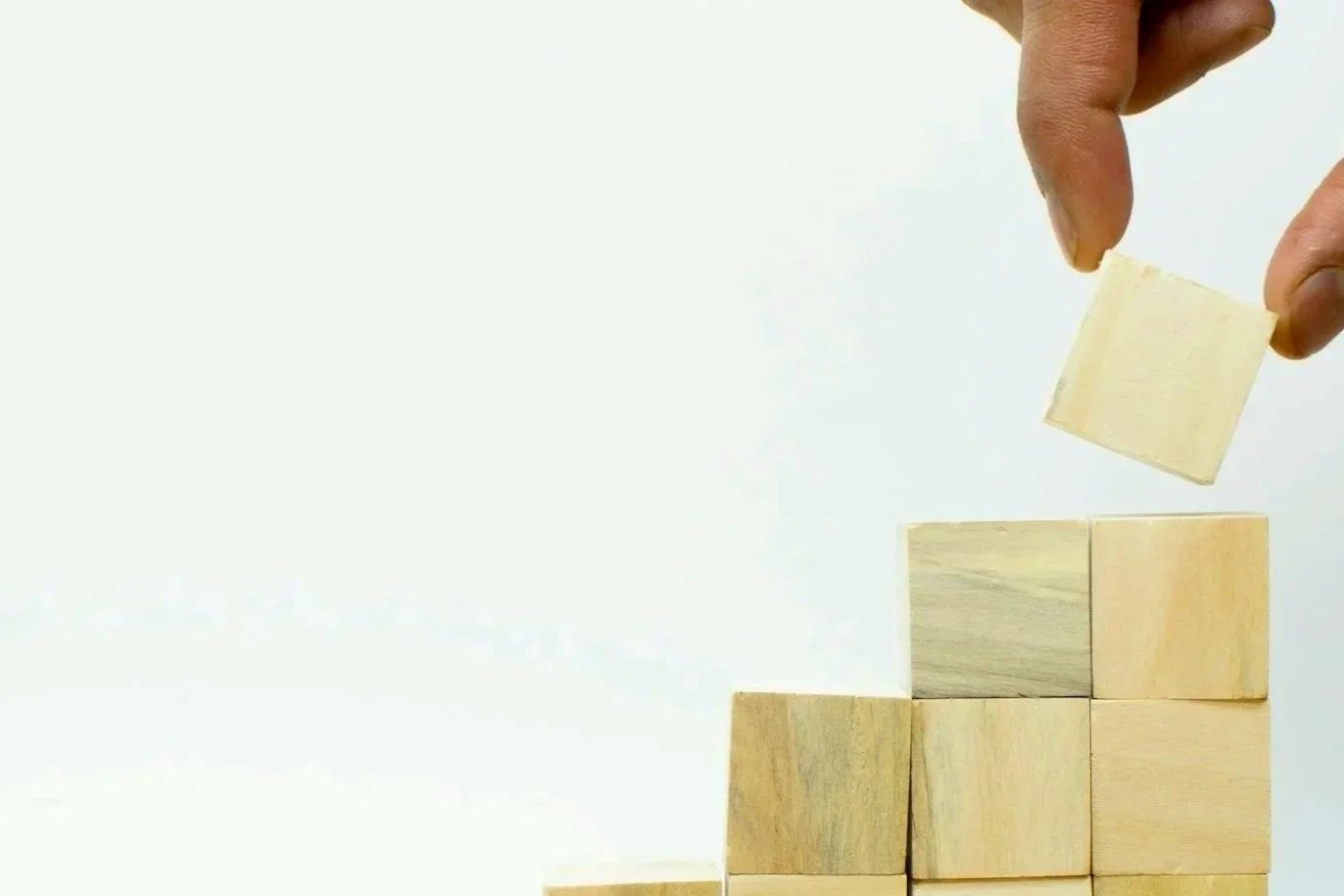Hand placing a wooden block on top of a stack of cubes against a plain background.