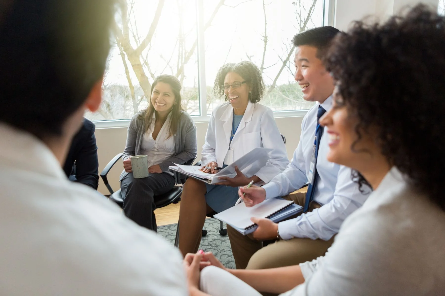 A diverse group of healthcare professionals, including doctors and nurses, engaged in a meeting and smiling in a bright conference room.
