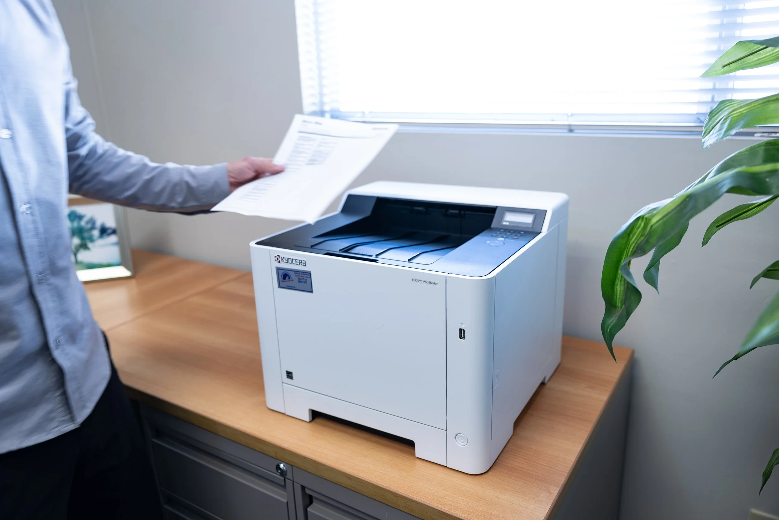A person in a light blue shirt is using a Kyocera monochrome laser printer to print a document, which they are holding in their left hand, on a wooden desk near a window with blinds.