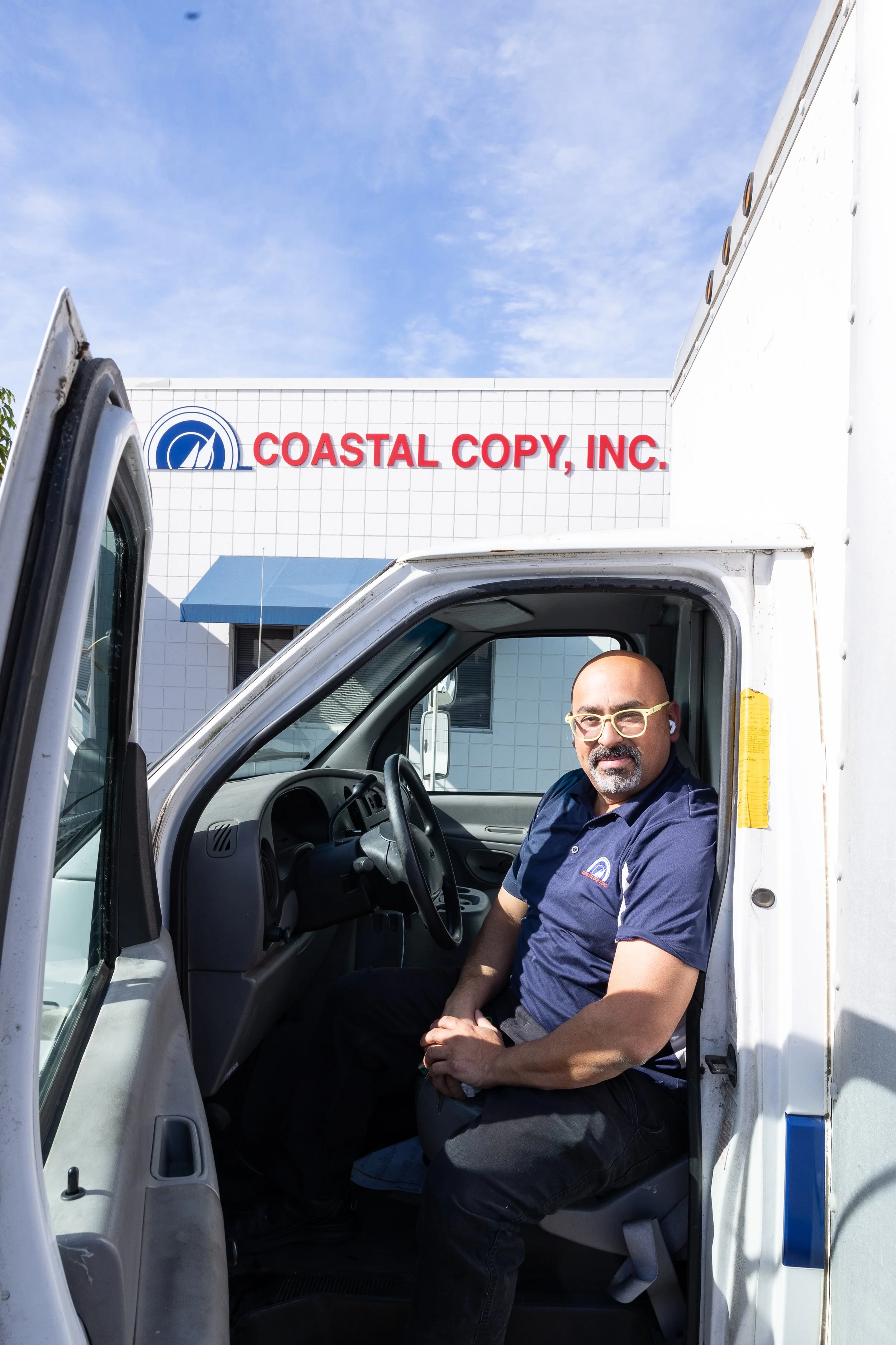 Man sitting in the driver's seat of a delivery truck parked outside a building with a sign that reads 'COASTAL COPY, INC.' under a blue sky.