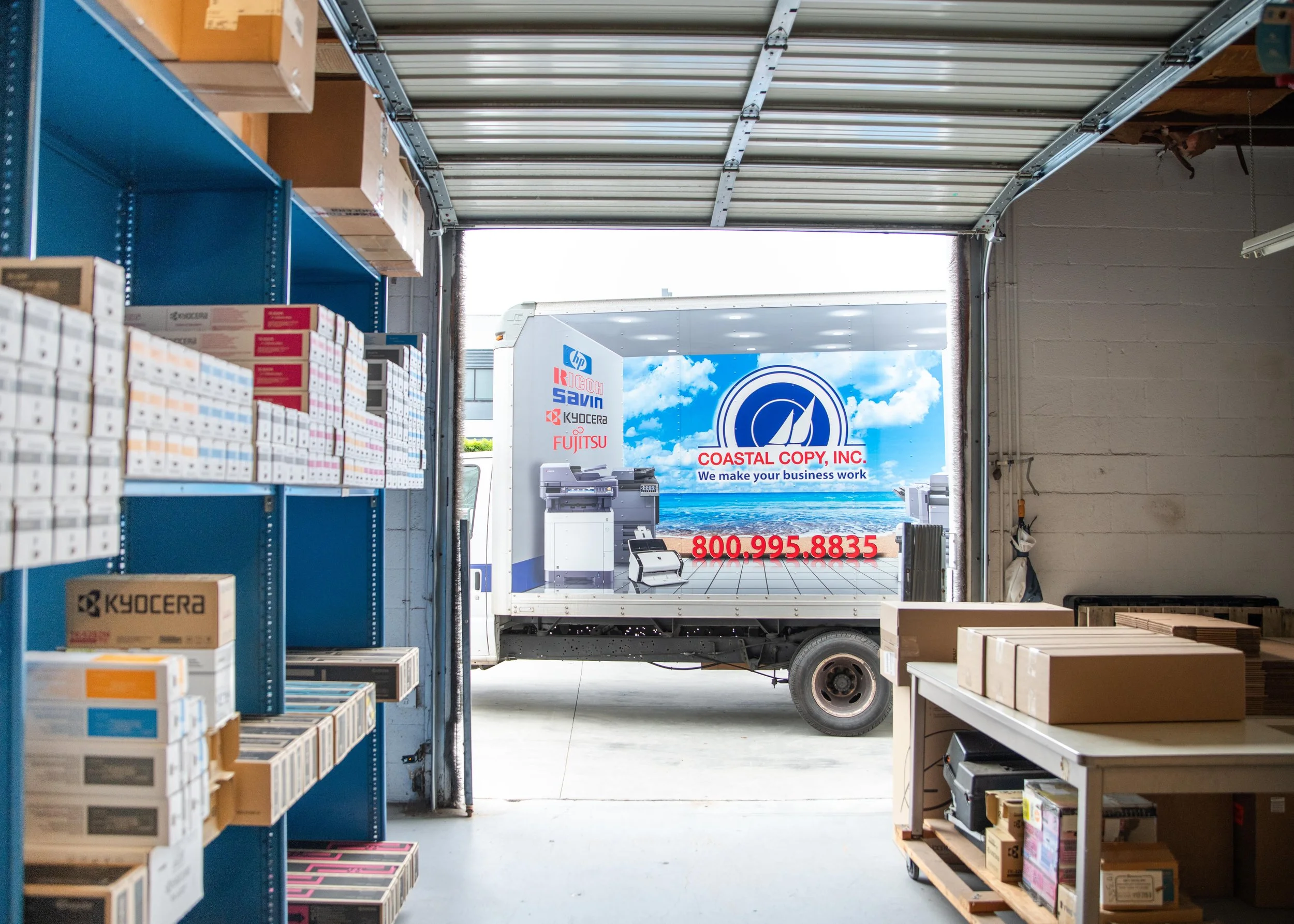 View inside a warehouse or storage facility with shelves holding boxes and a table, looking out through a garage door to a delivery truck outside with advertising for Coastal Copy, Inc., including a sea and sky background, company logo, phone number, and images of office equipment.