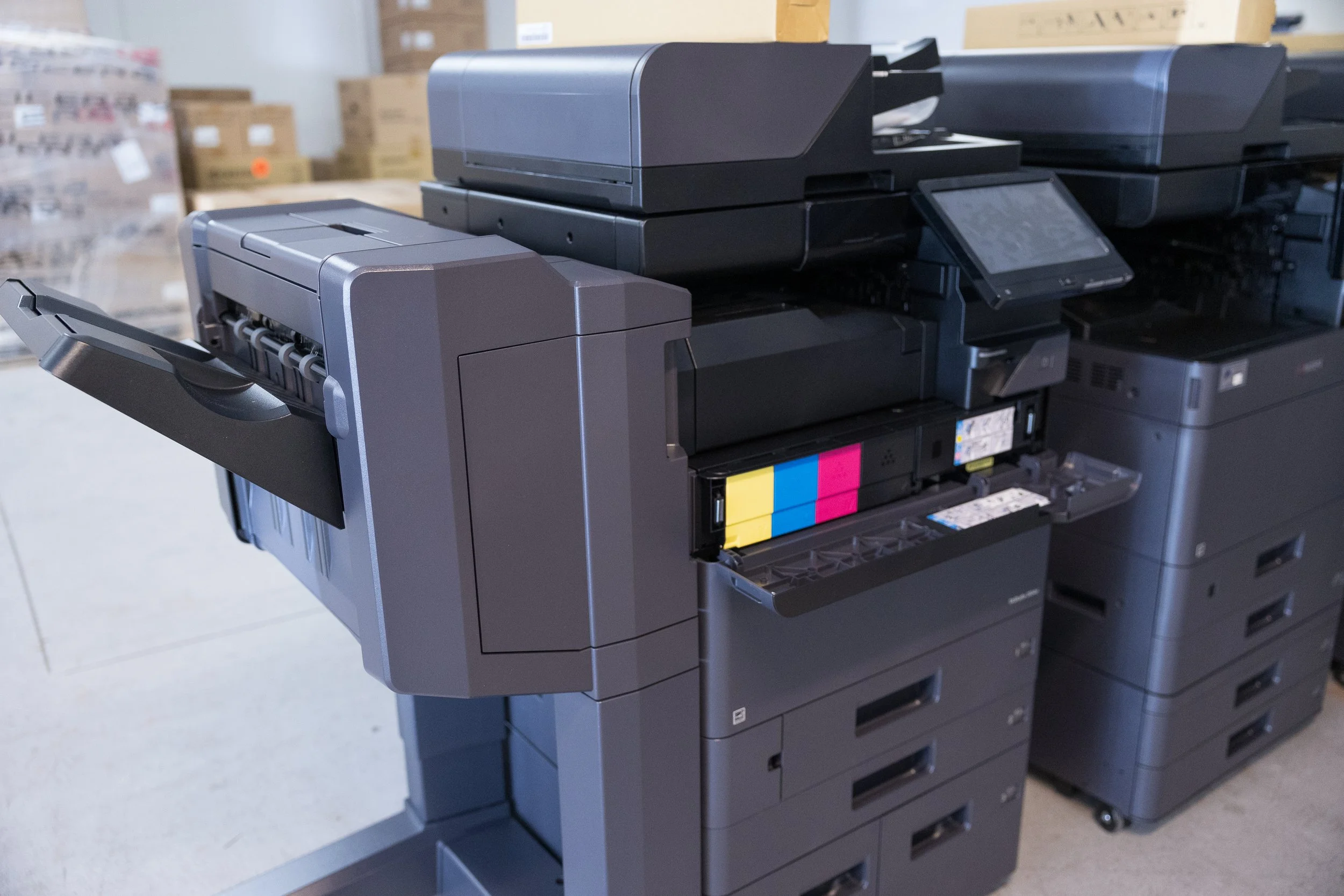 A black multifunction copier with a paper tray, situated in a warehouse, with cardboard boxes in the background.