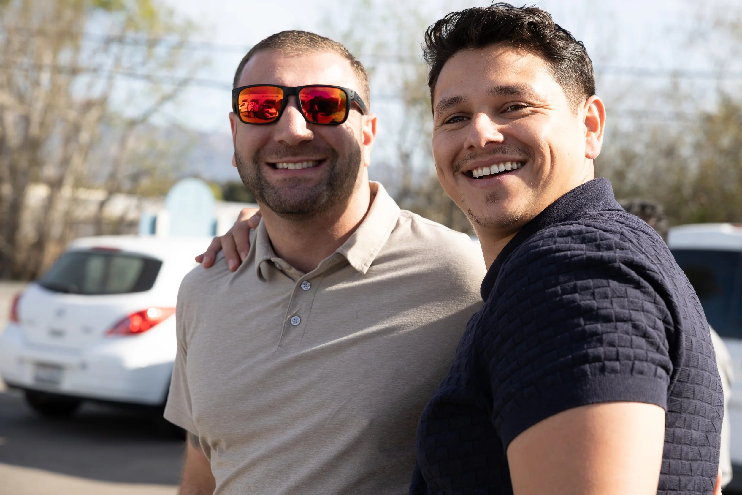 Two smiling men standing outdoors, one wearing sunglasses and a beige polo shirt, the other in a dark blue textured shirt, with cars and trees in the background.