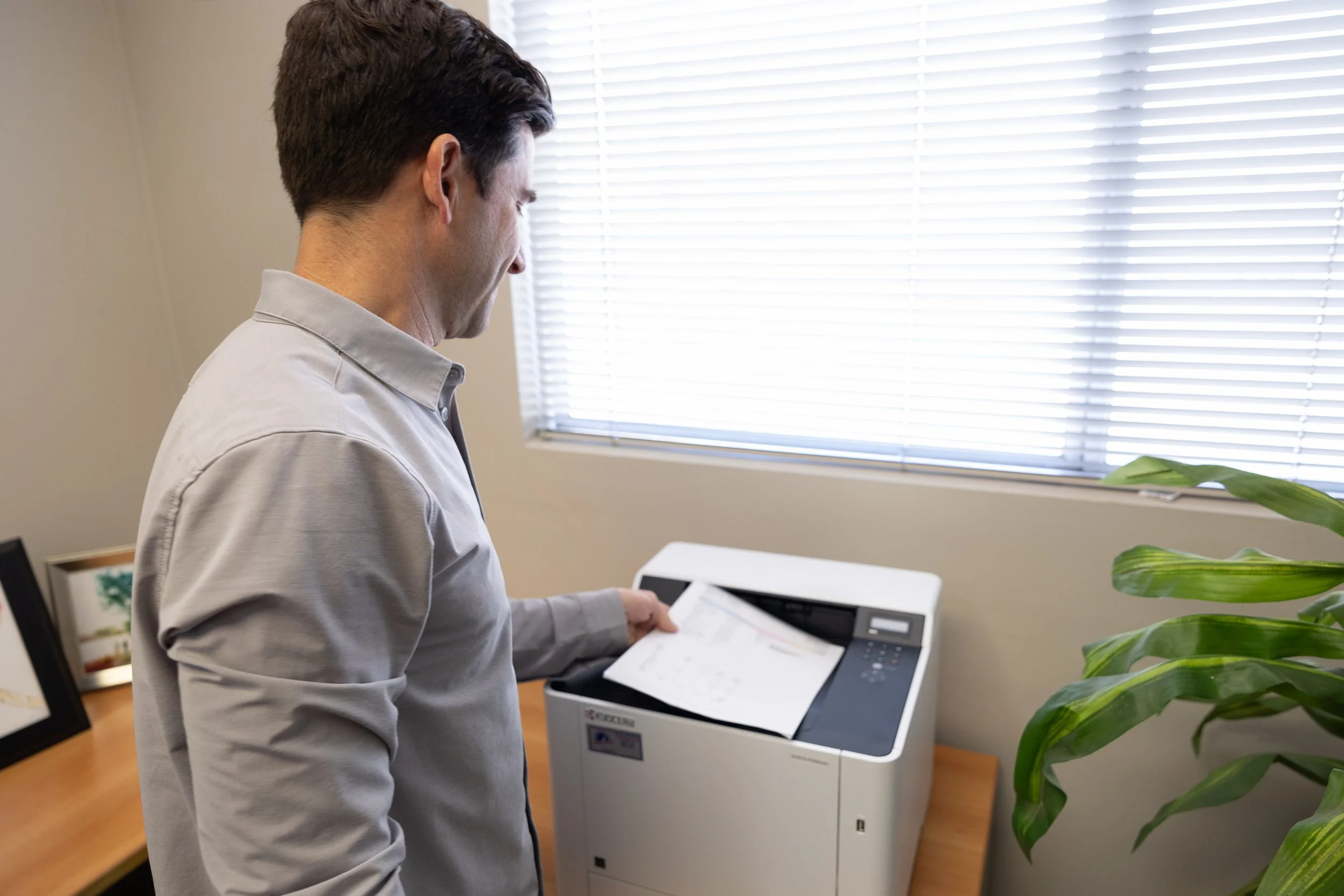 A man in a light gray shirt using a printer with paper inside, near a window with blinds in an office.